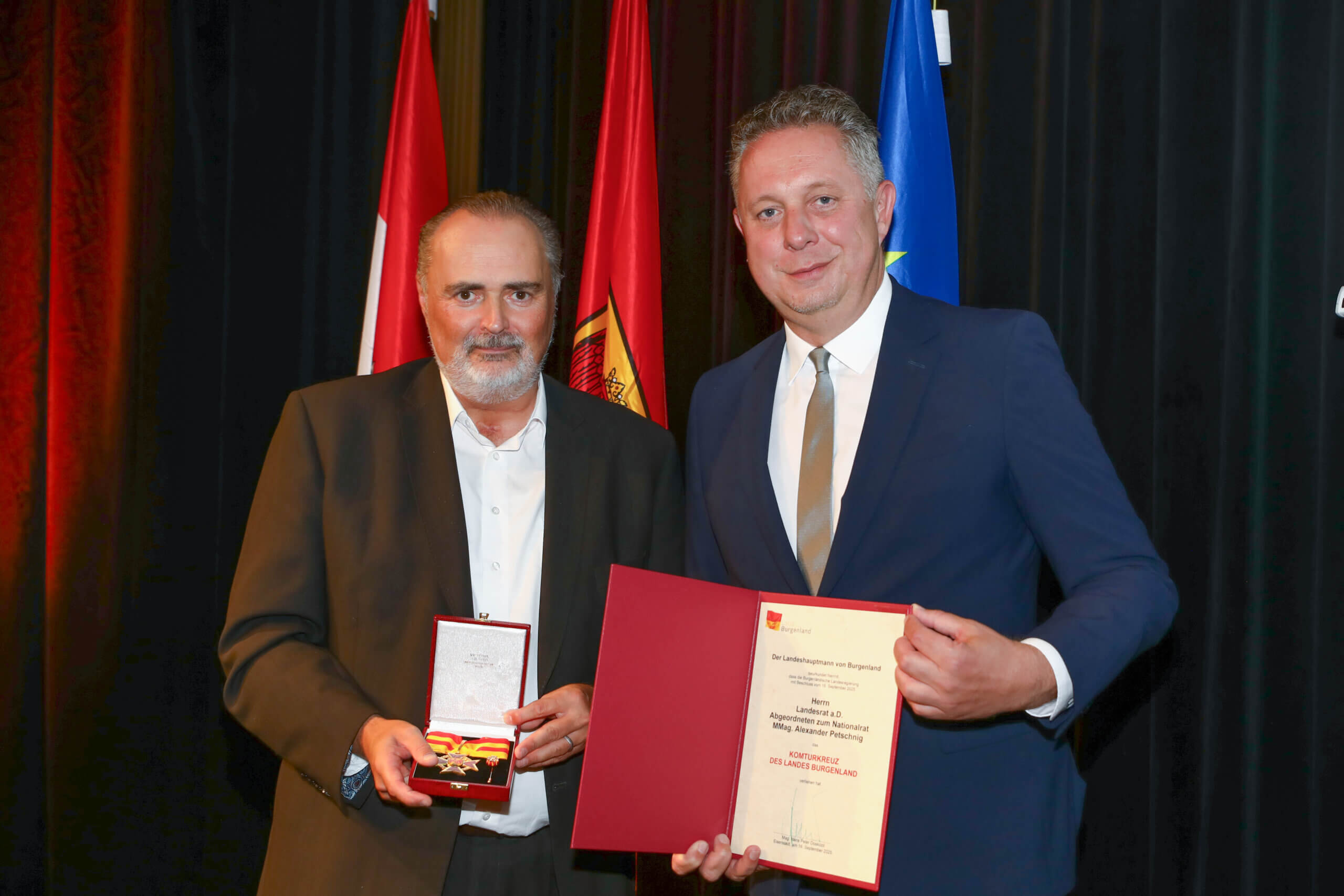 Zwei Männer bei Preisverleihung mit Urkunde und Medaille, vor farbigen Fahnen auf der Bühne.