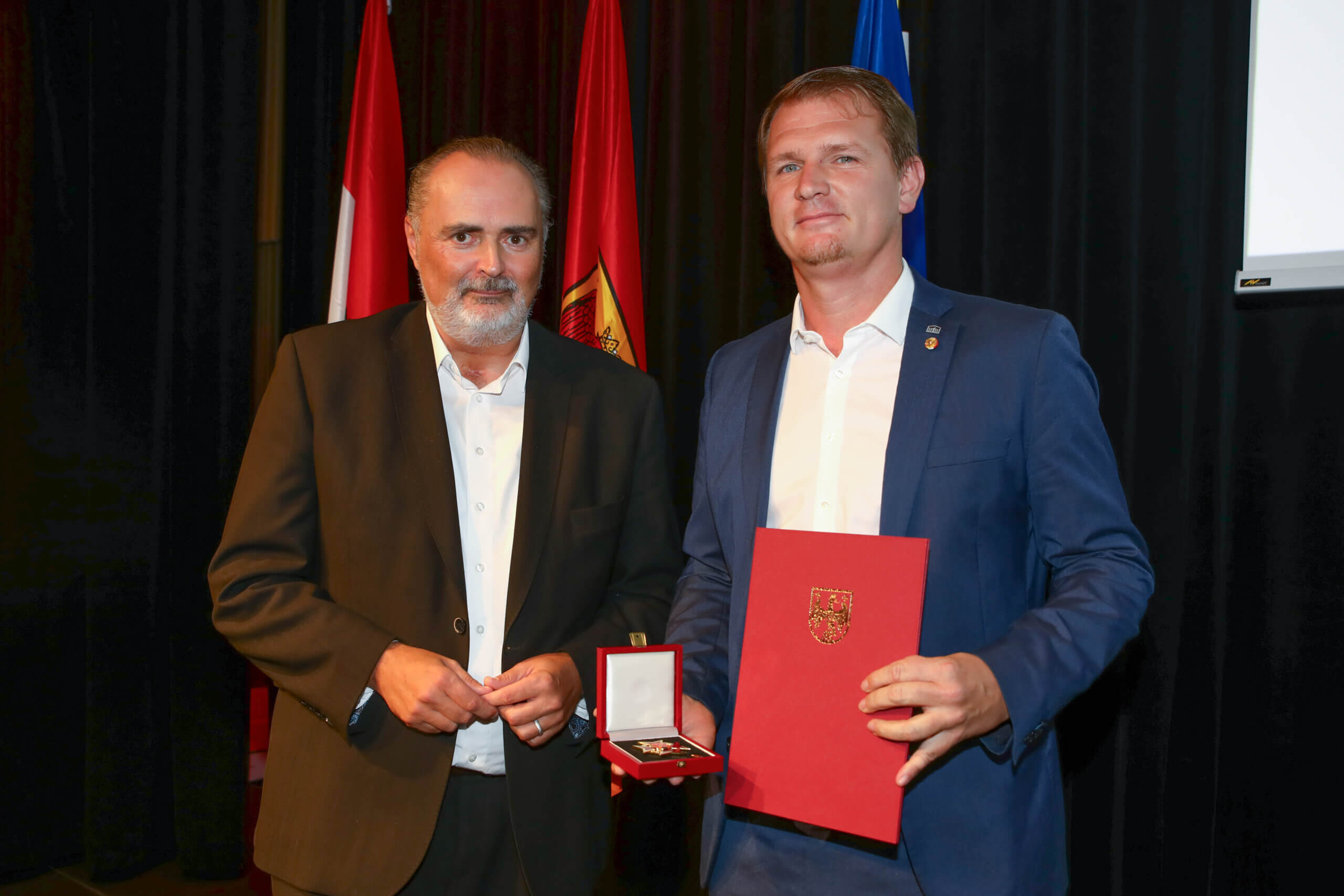 Zwei Männer bei einer Ehrung mit Urkunde und Medaille vor Fahnen auf einer Bühne.