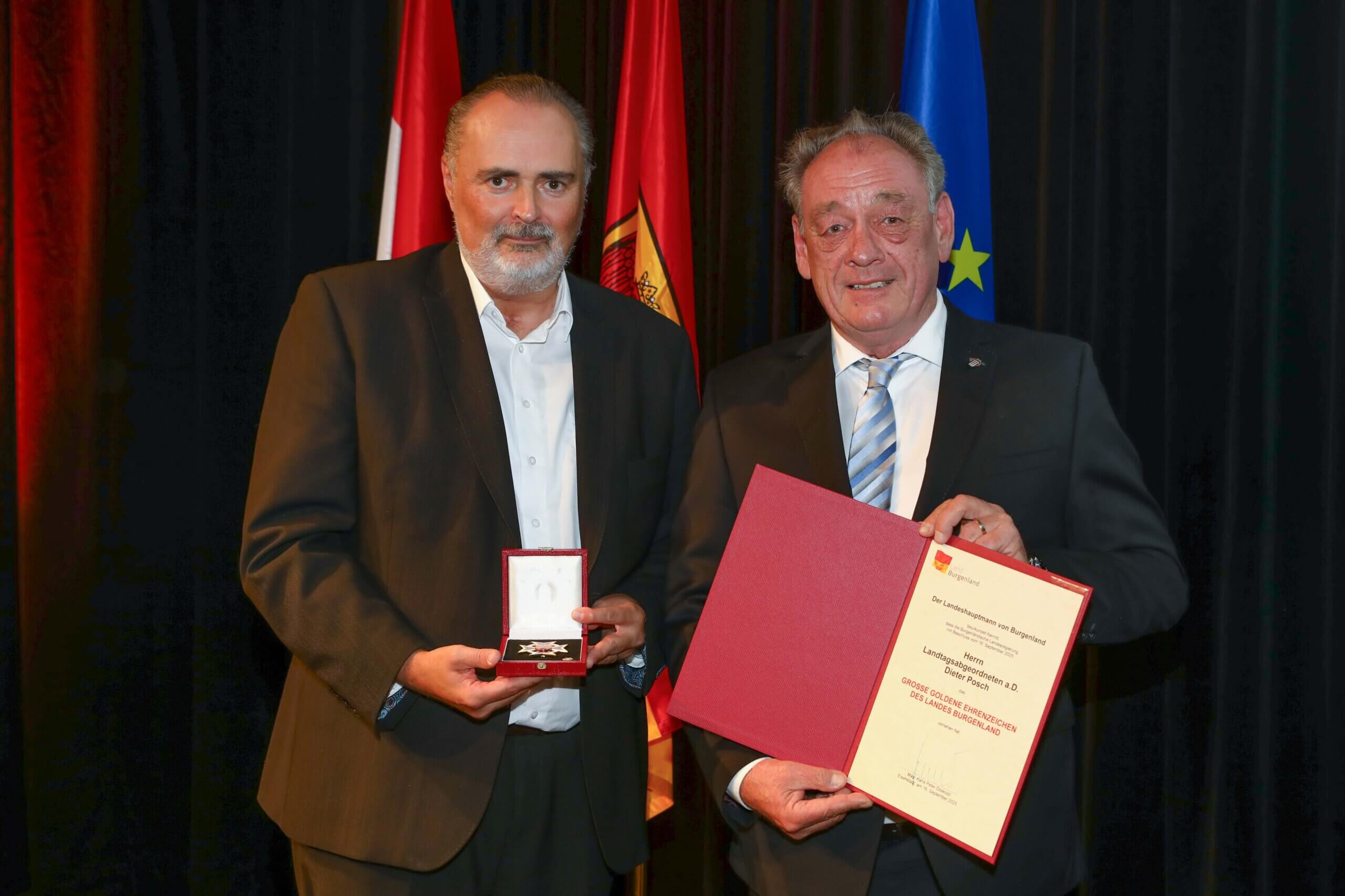 Zwei Männer bei einer Preisverleihung, einer hält eine Auszeichnung, der andere ein Zertifikat vor mehreren Flaggen.