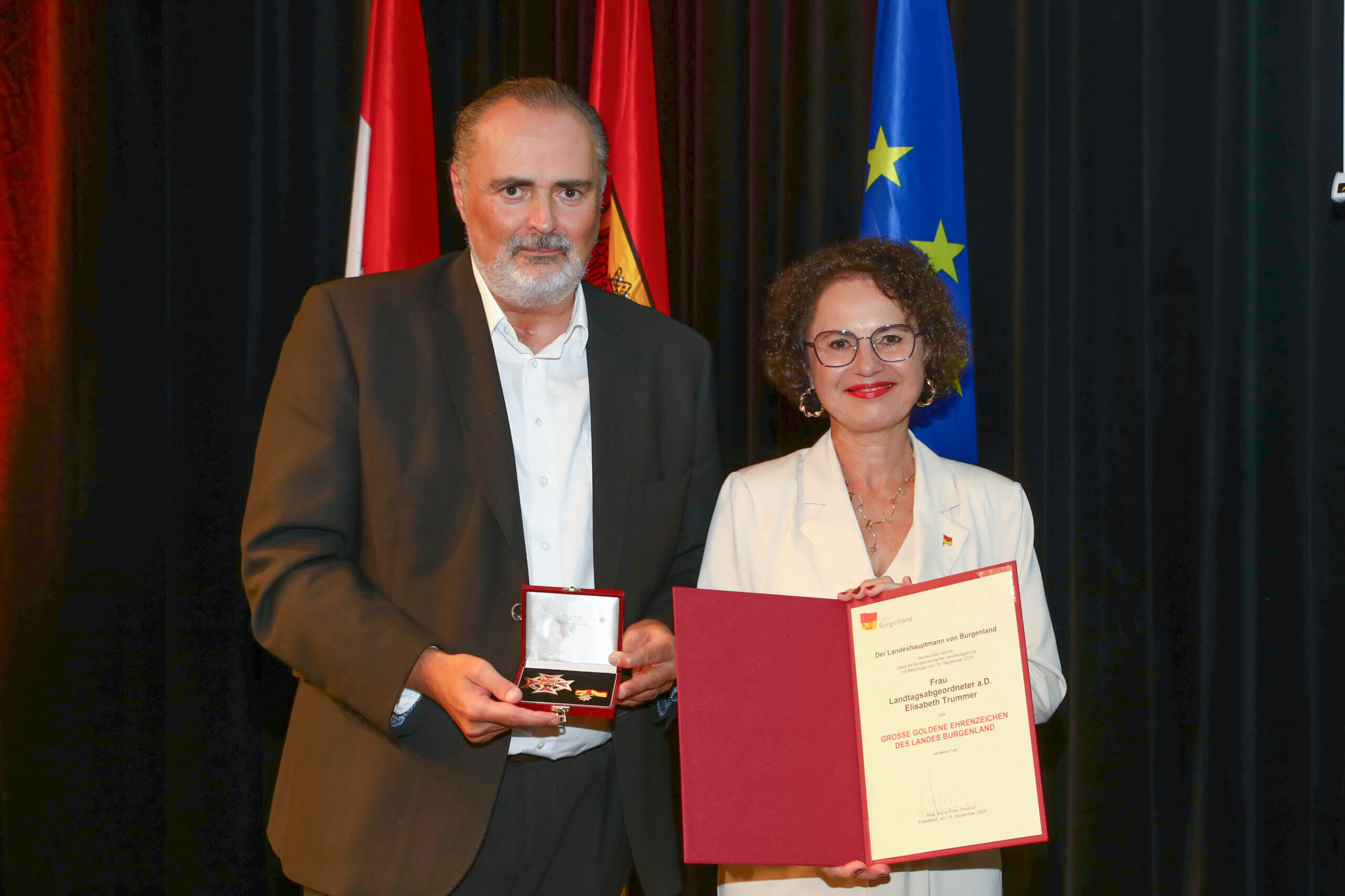 Person erhält Auszeichnung und Urkunde, stehen vor Fahnen und lächeln. Ehrung im festlichen Rahmen.
