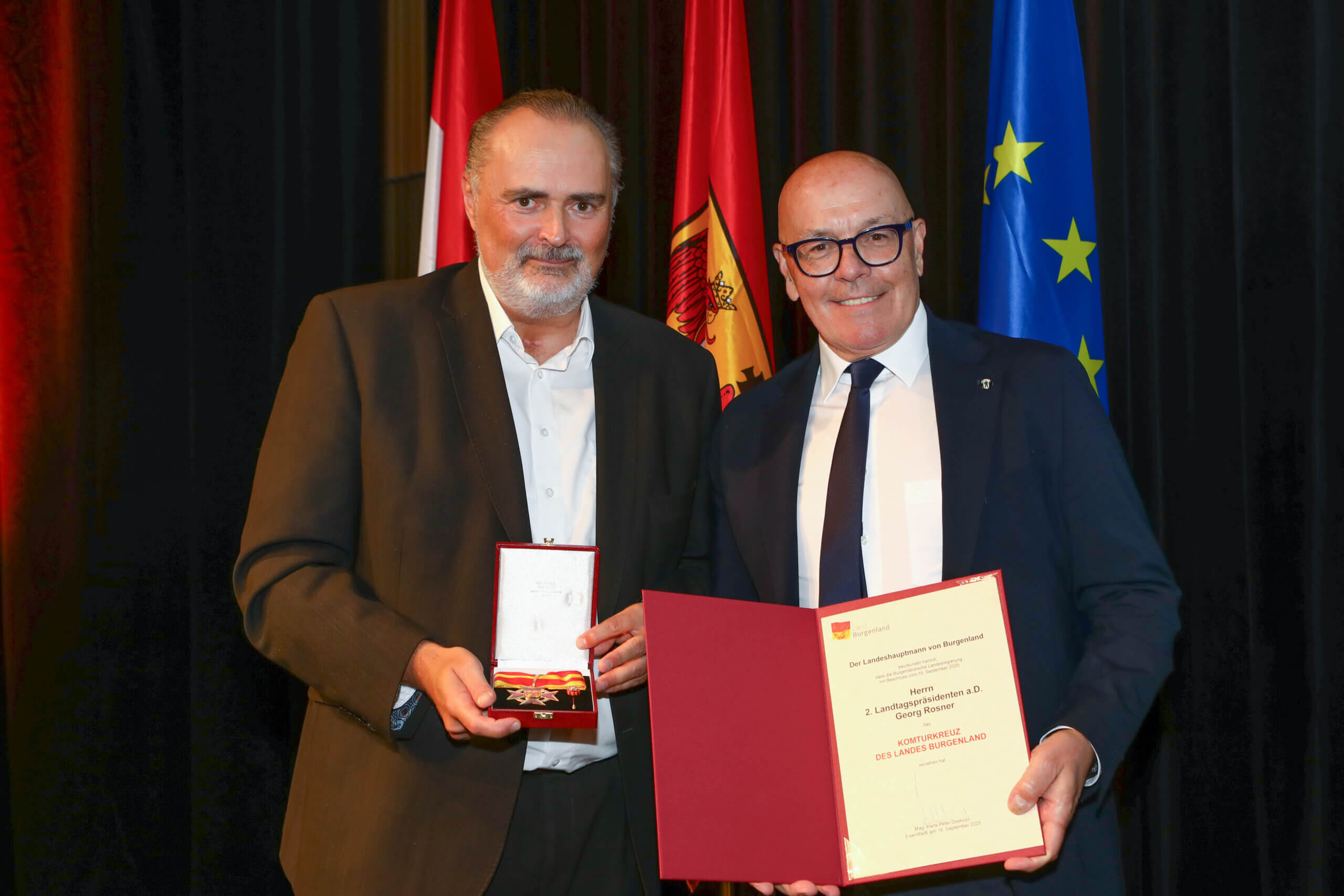 Männer bei Preisverleihung vor Flaggen; einer hält eine Medaille, der andere zeigt eine Urkunde.