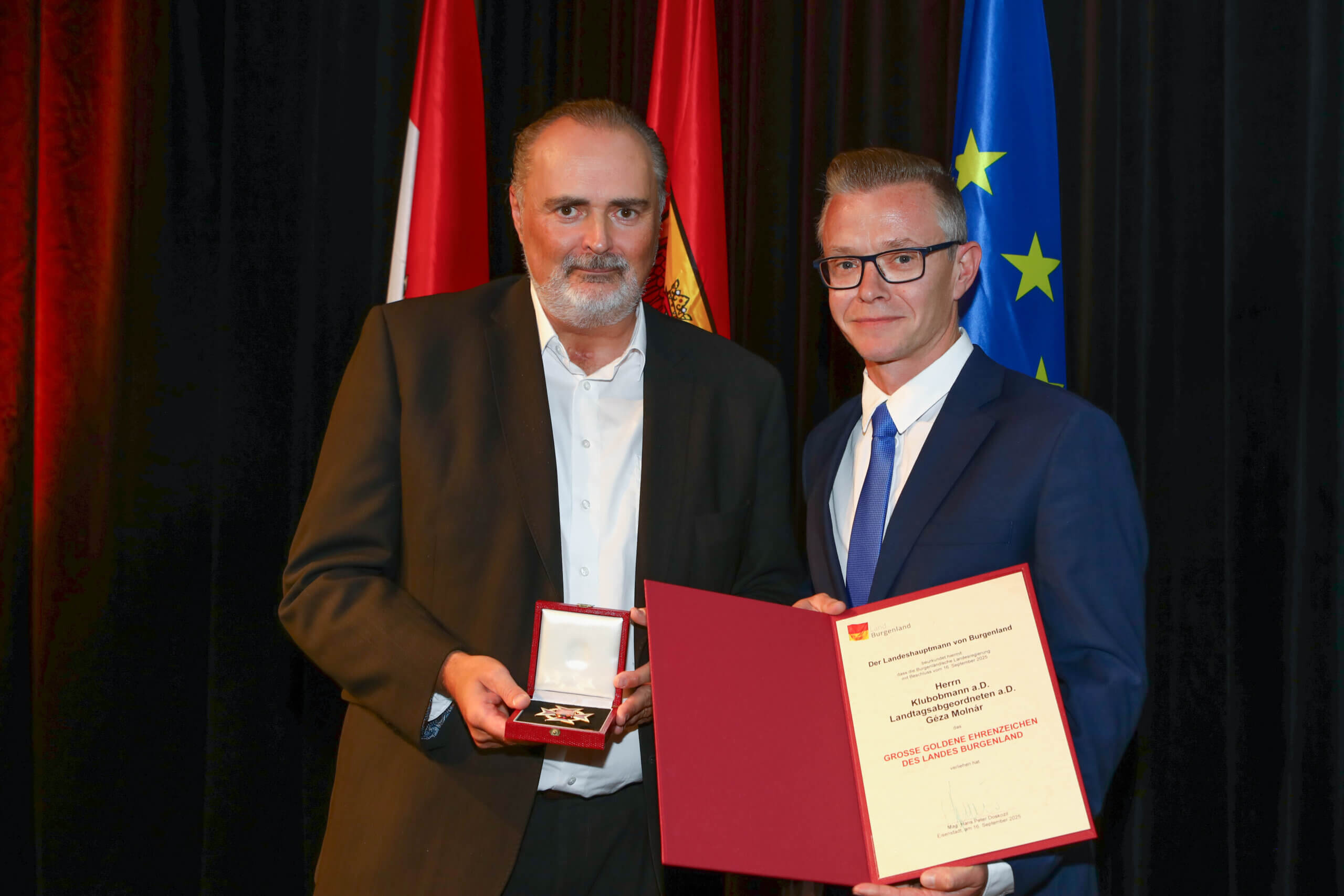 Zwei Männer bei einer Ehrung mit Auszeichnung und Urkunde, vor Flaggen in einem formellen Setting.