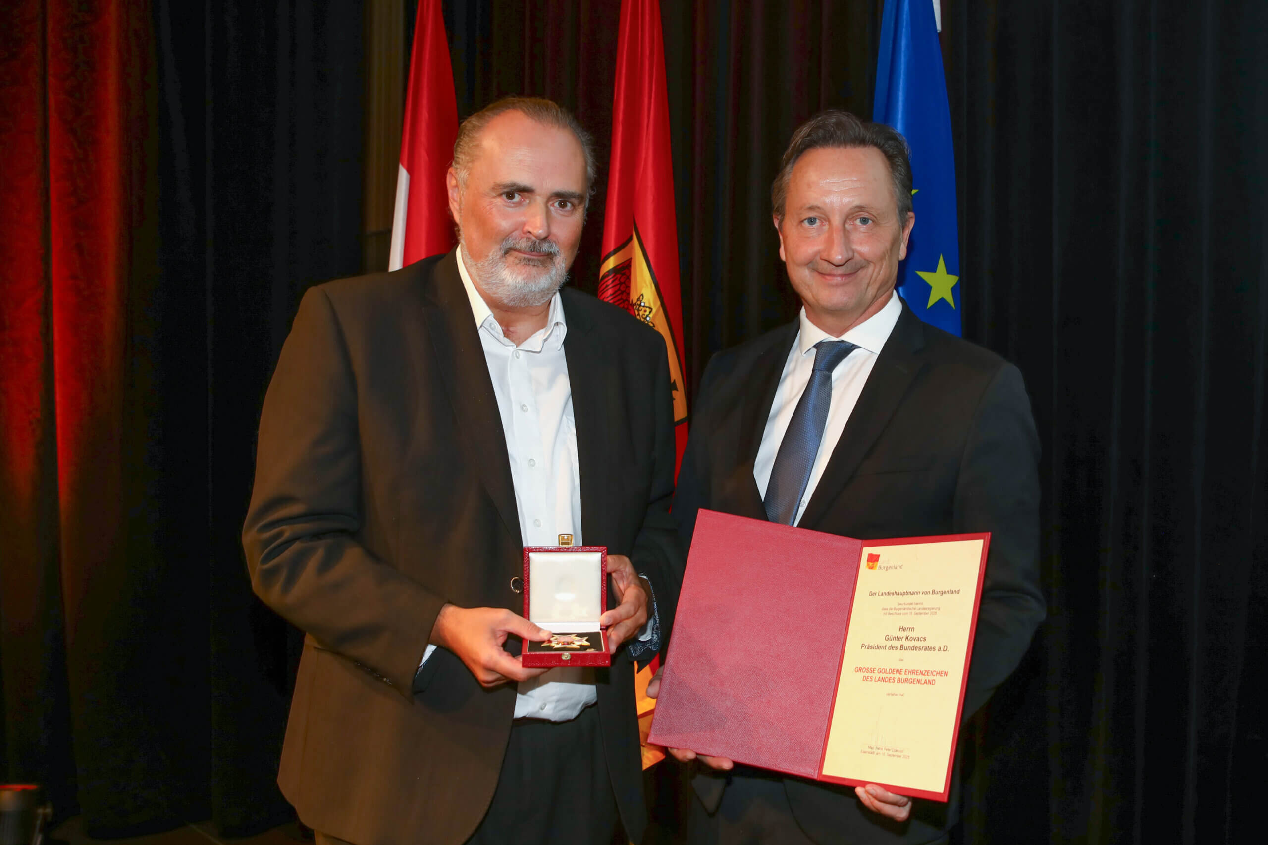 Zwei Männer mit Auszeichnung und Urkunde bei einer Preisverleihung, europäische Flagge im Hintergrund.
