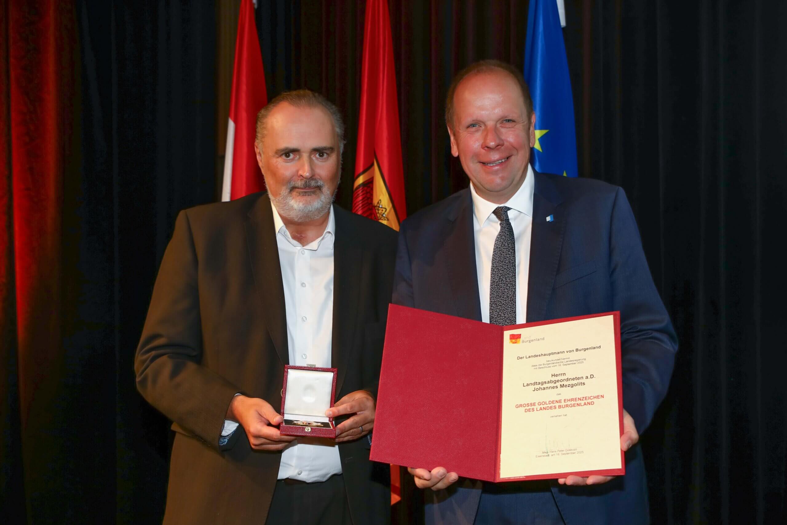 Zwei Männer bei der Verleihung einer Auszeichnung in Burgenland vor Flaggen, einer hält eine Medaille, der andere eine Urkunde.