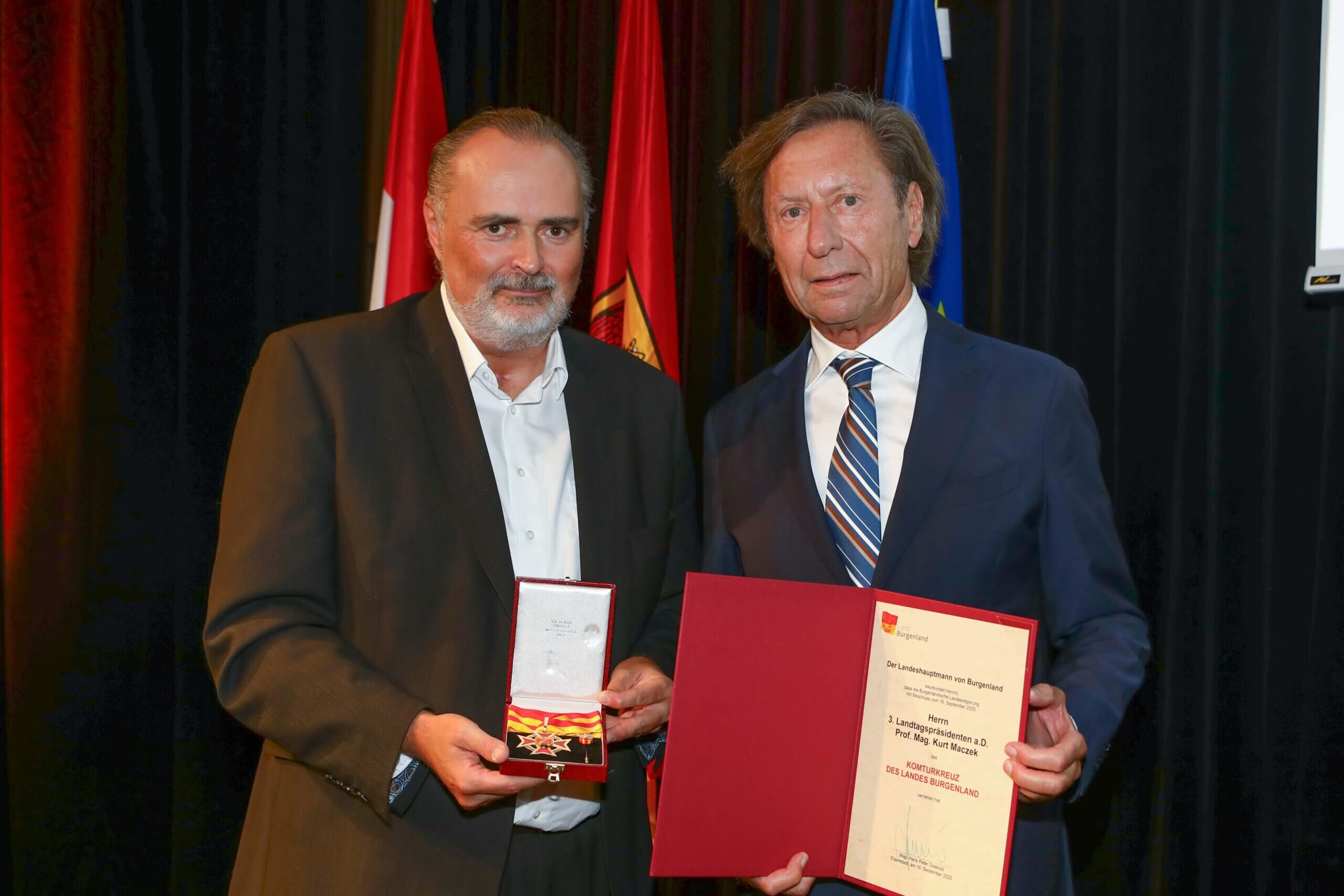 Zwei Männer mit Urkunde und Medaille bei einer Auszeichnung in einem feierlichen Rahmen, mit Flaggen im Hintergrund.