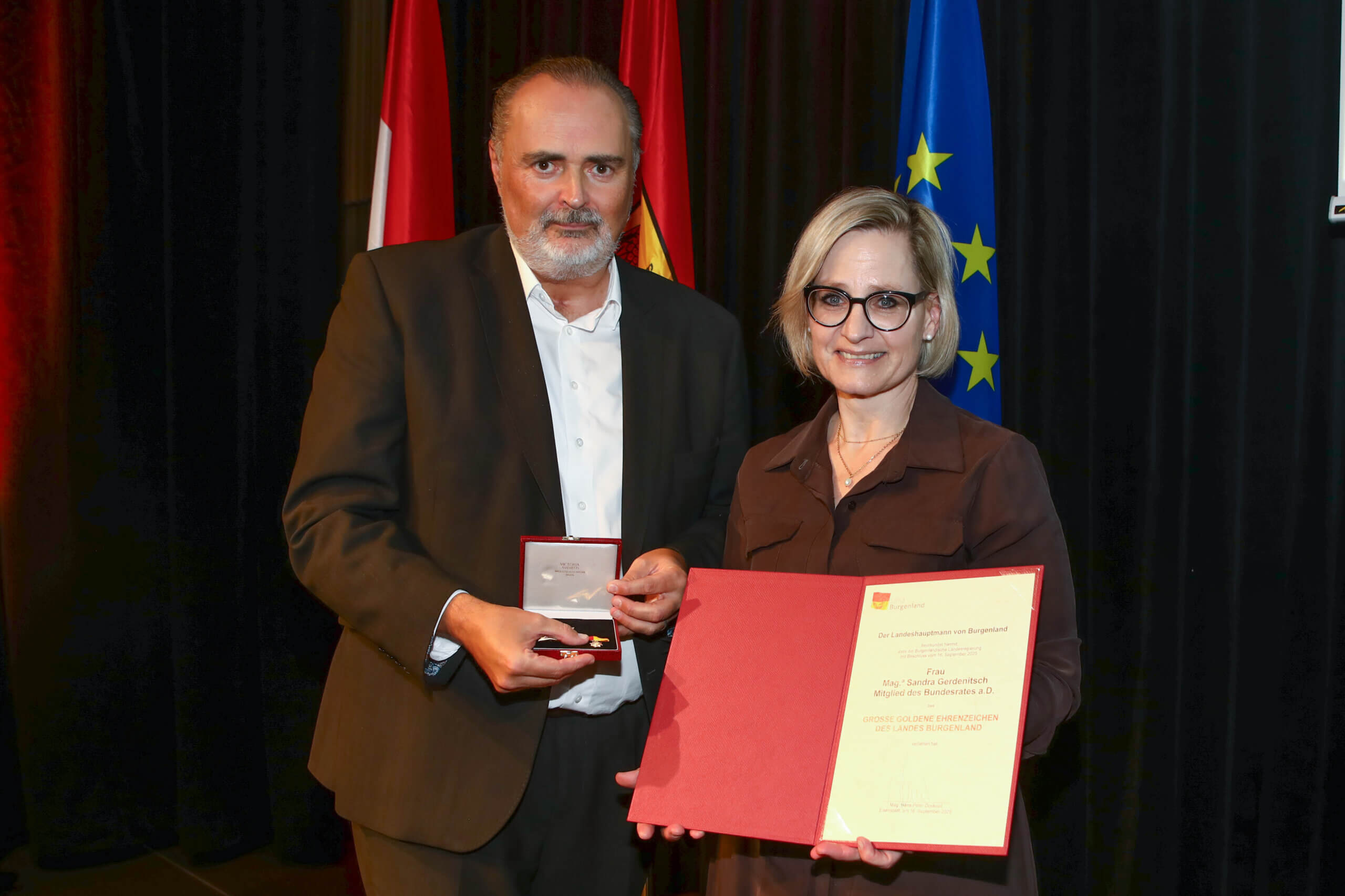 Personen halten eine Auszeichnung und Urkunde vor Flaggen bei einer feierlichen Zeremonie.