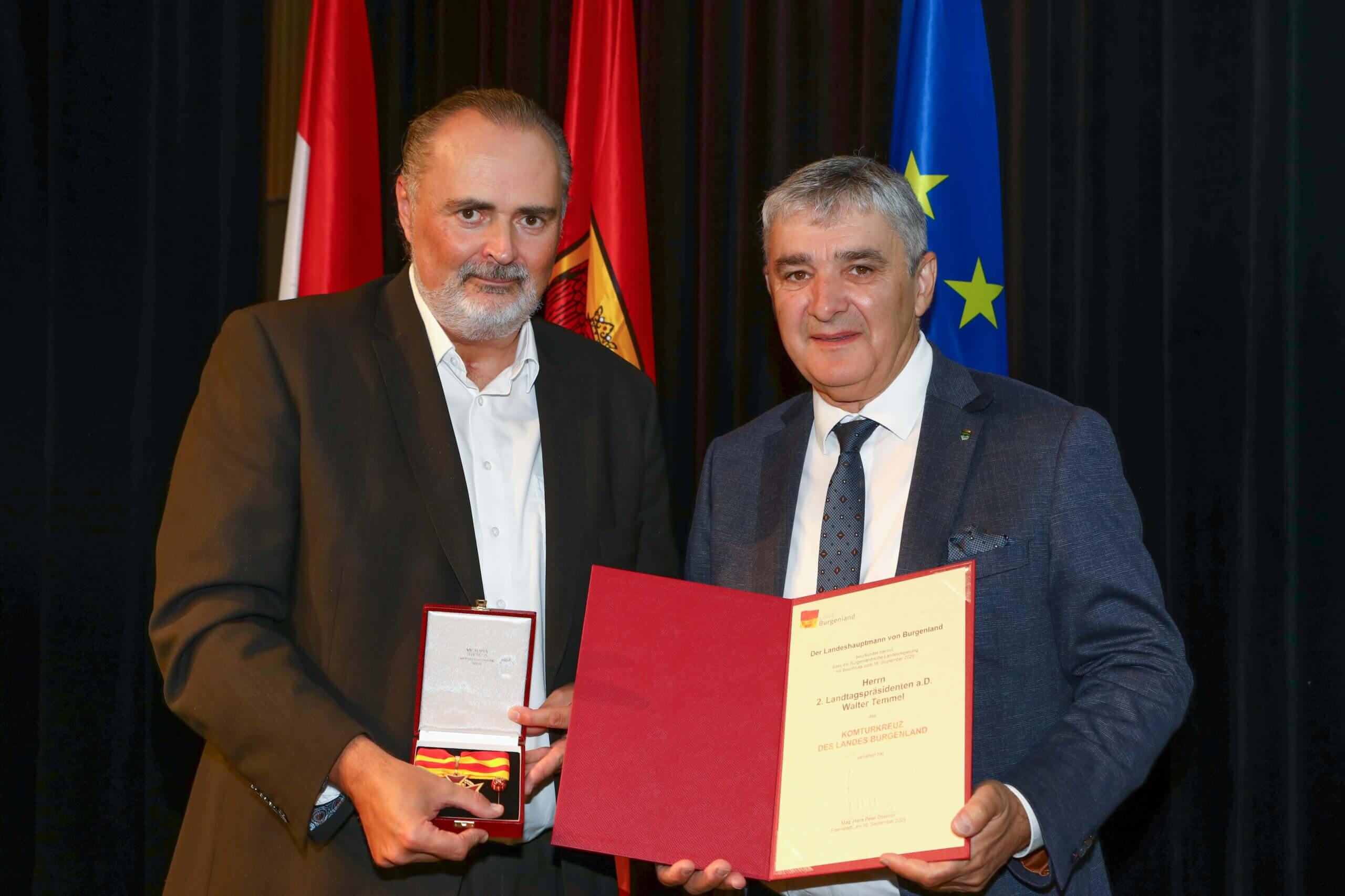 Zwei Männer bei einer Auszeichnungszeremonie, einer hält eine Medaille und ein Zertifikat vor Flaggen im Hintergrund.