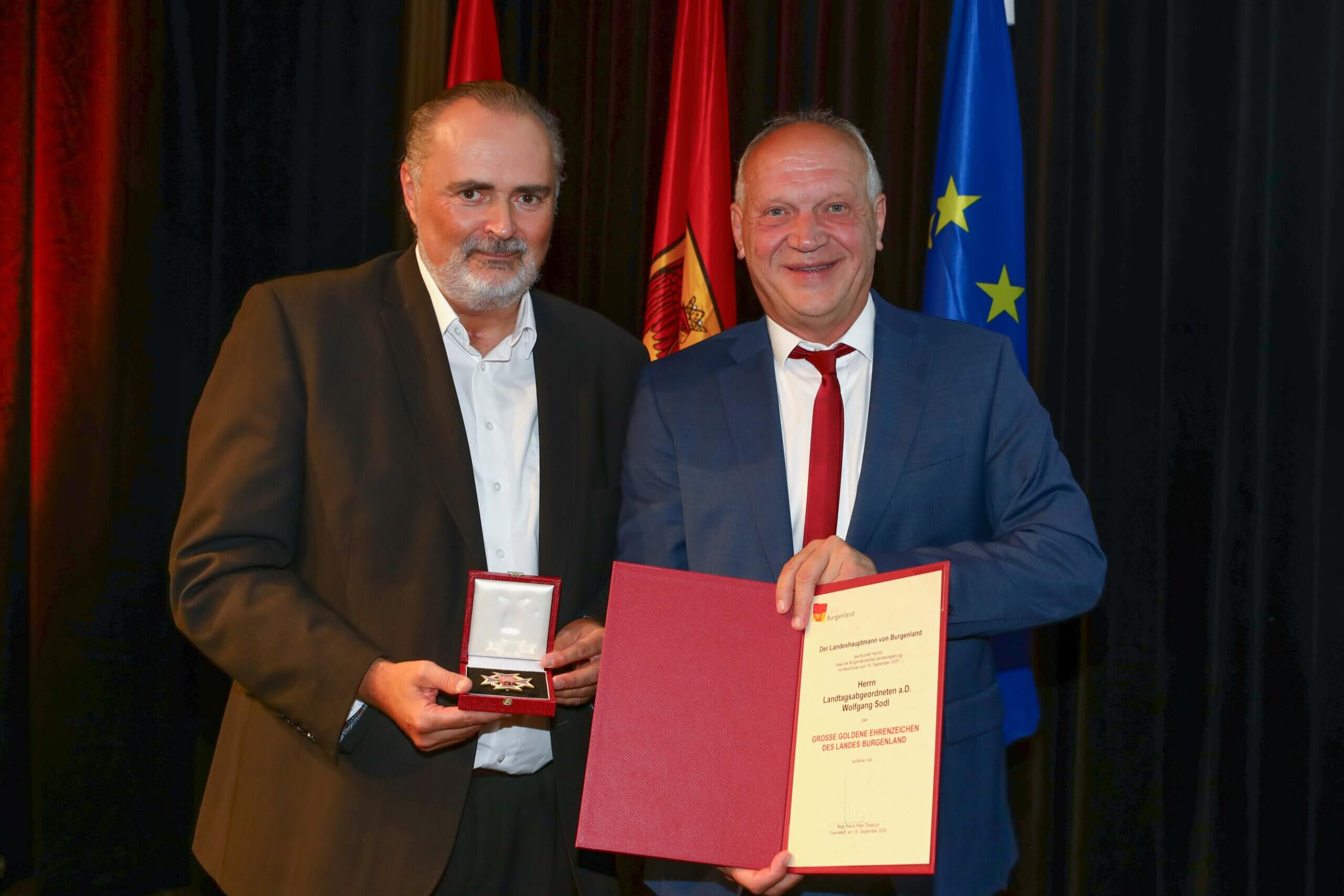 Zwei Männer bei einer Preisverleihung im Burgenland mit Urkunde und Medaille, vor nationalen und EU-Flaggen.