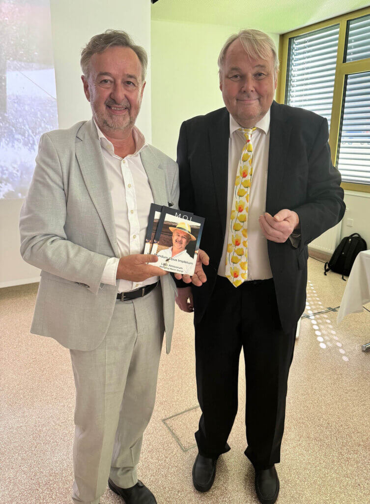 Zwei Herren bei einer Buchpräsentation, einer hält ein Buch mit einem Coverbild in die Kamera, beide tragen Anzüge.