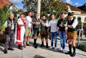 Gruppe von Männern im Freien mit traditionellen Trachten, die gemeinsam auf einem Holzdeck anstoßen.