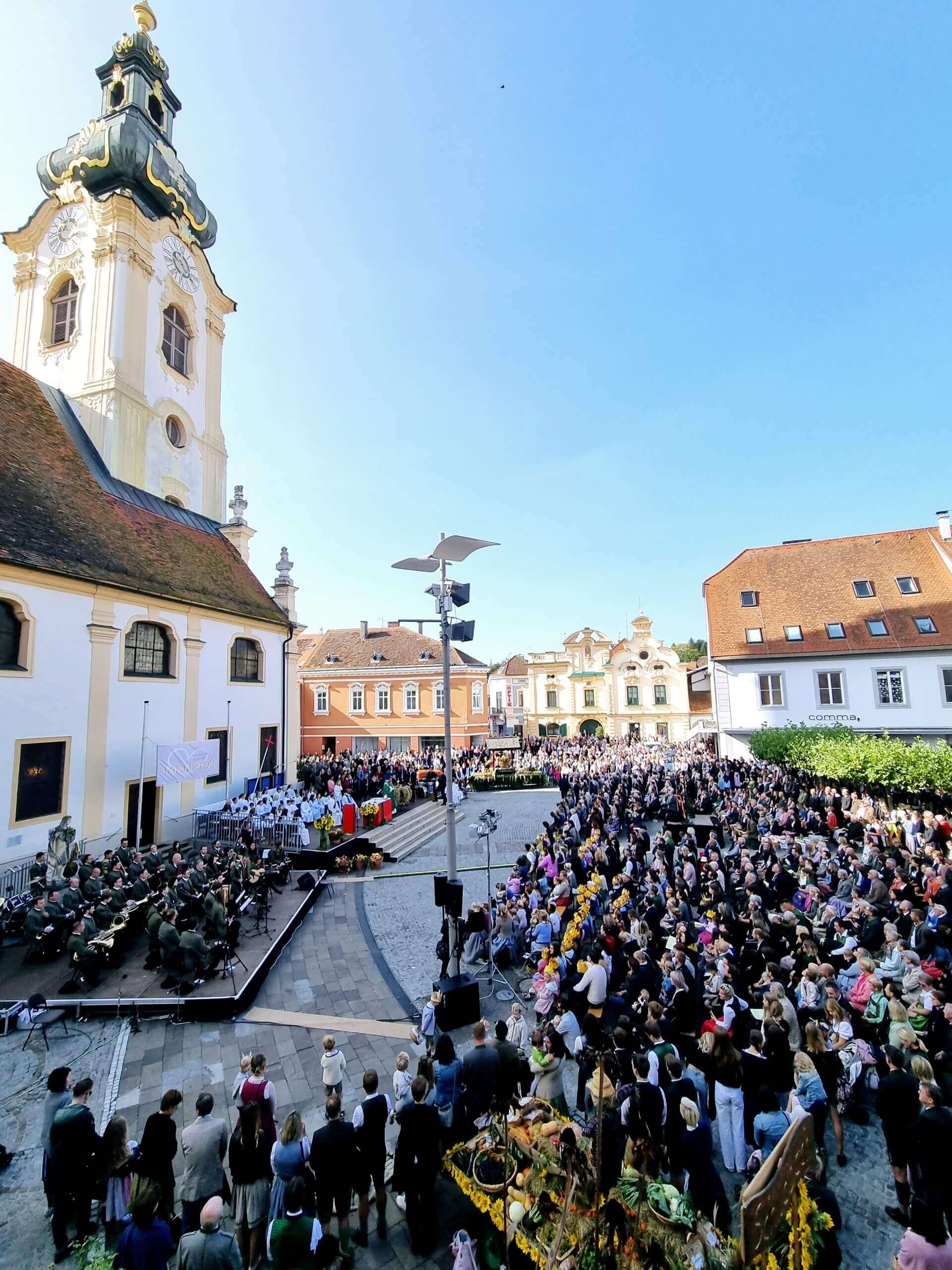 Menschenmenge bei einem Kirchenfest vor barocker Kirche unter strahlend blauem Himmel, dekoriert mit Blumen und Erntegaben.