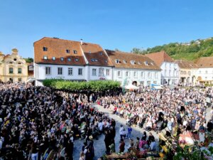 Große Menschenmenge versammelt sich bei klarem Himmel auf einem Platz vor historischen Gebäuden für ein lokales Fest.