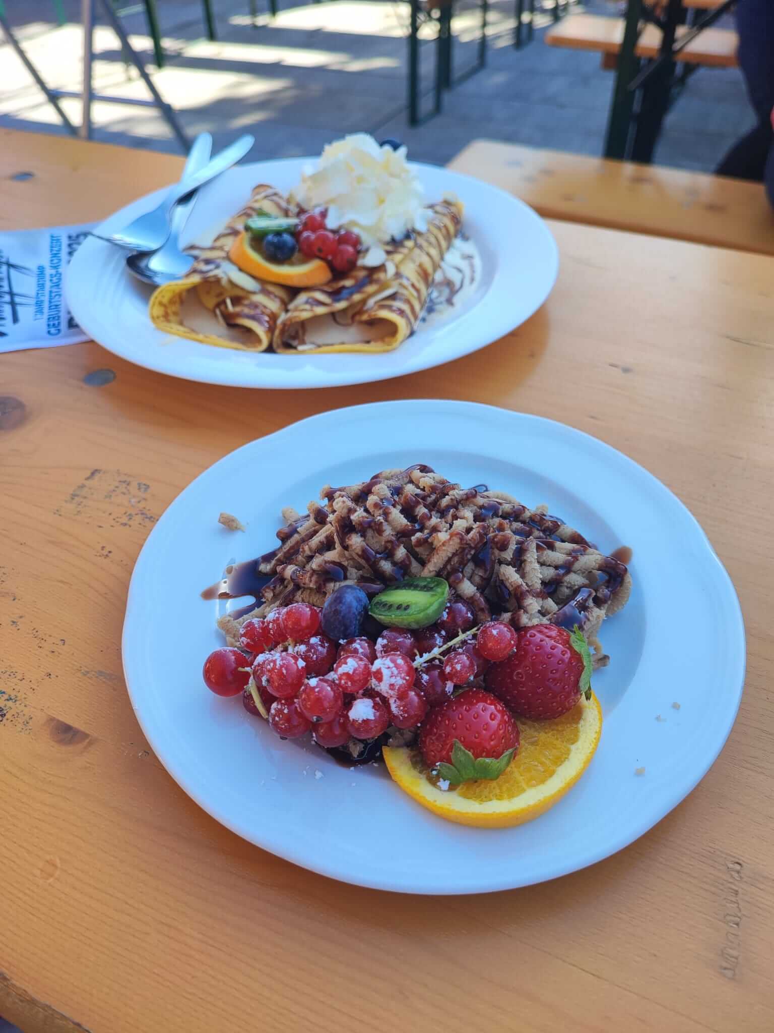 Leckere Dessertplatten mit Crepes, Sahne, Schokosoße, Beeren und Waffeln auf einem Holztisch im Freien.