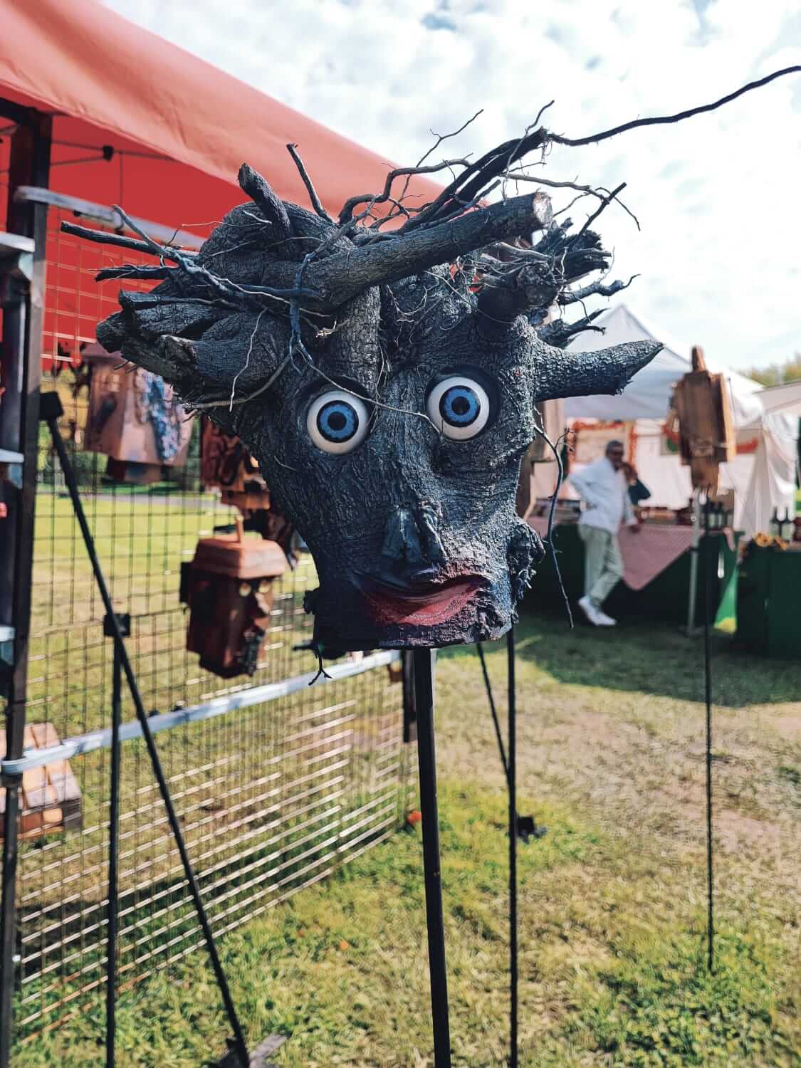 Skulptur aus Zweigen mit großen Augen auf einem Kunsthandwerkermarkt, umgeben von Ständen und Sonnenschirmen.