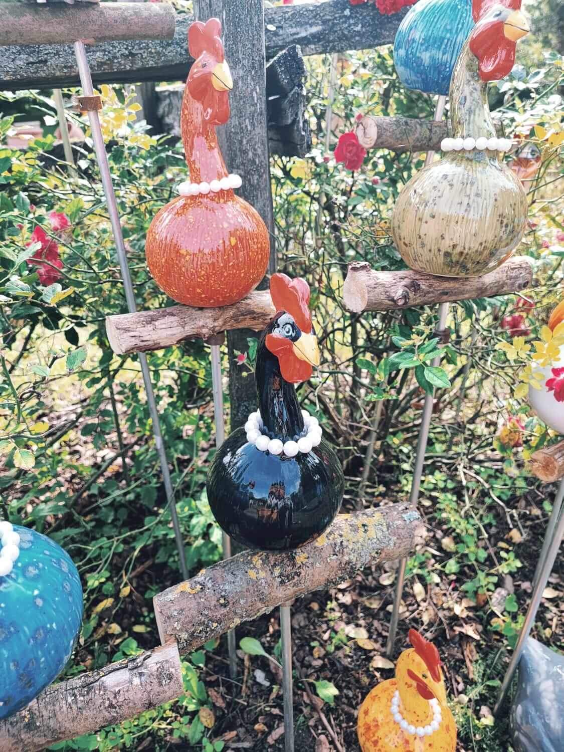 Bunte Keramik-Hühner auf Zweigen in einem Garten, dekorativ und handgefertigt, ideal für eine kreative Gartengestaltung.