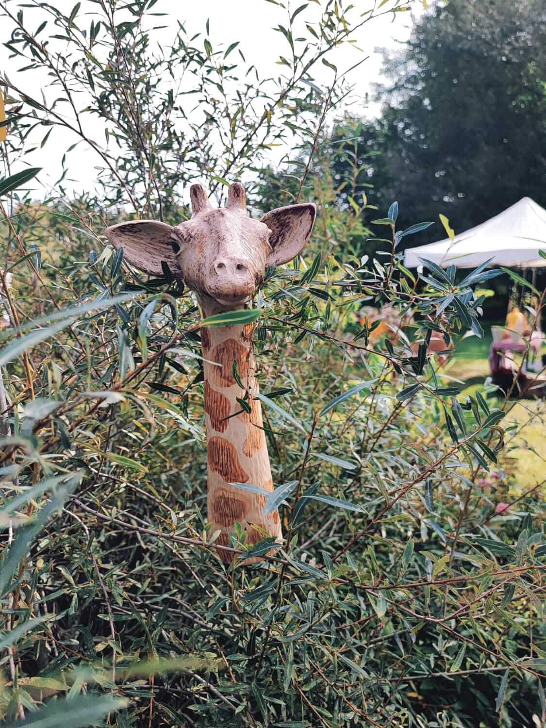 Holzgiraffen-Skulptur im Garten, umgeben von grünen Büschen, neben einem weißen Zelt bei Tageslicht.