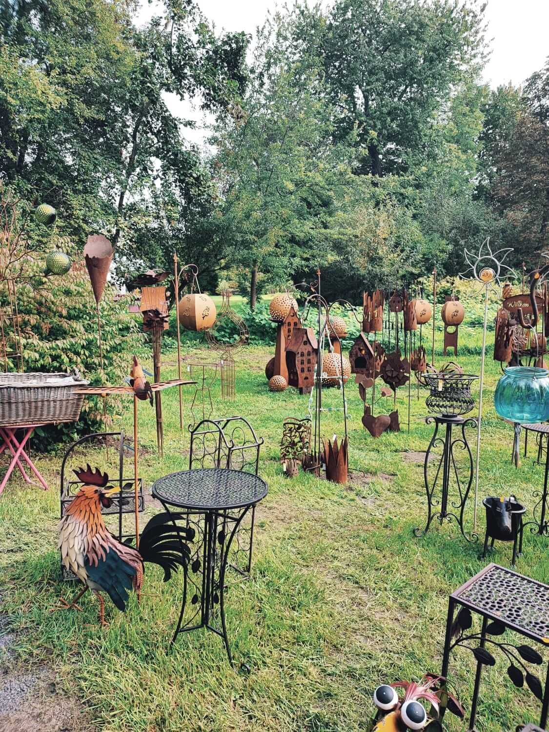 Garten voller Metallskulpturen: Tische, Stühle und Deko-Objekte aus Metall auf grüner Wiese umgeben von Bäumen.