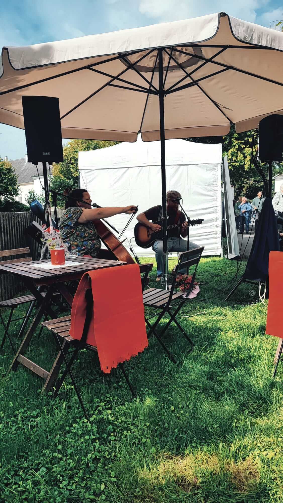 Zwei Musiker spielen Gitarre und Cello in einem Garten unter einem großen Sonnenschirm bei einem Freiluft-Event.