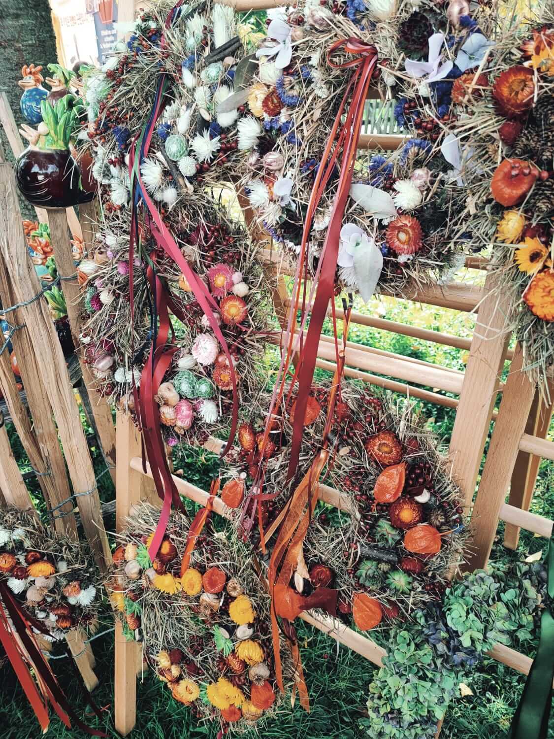Herbstliche Türkränze aus Trockenblumen und bunten Bändern an einem Holzgestell auf einem Markt ausgestellt.