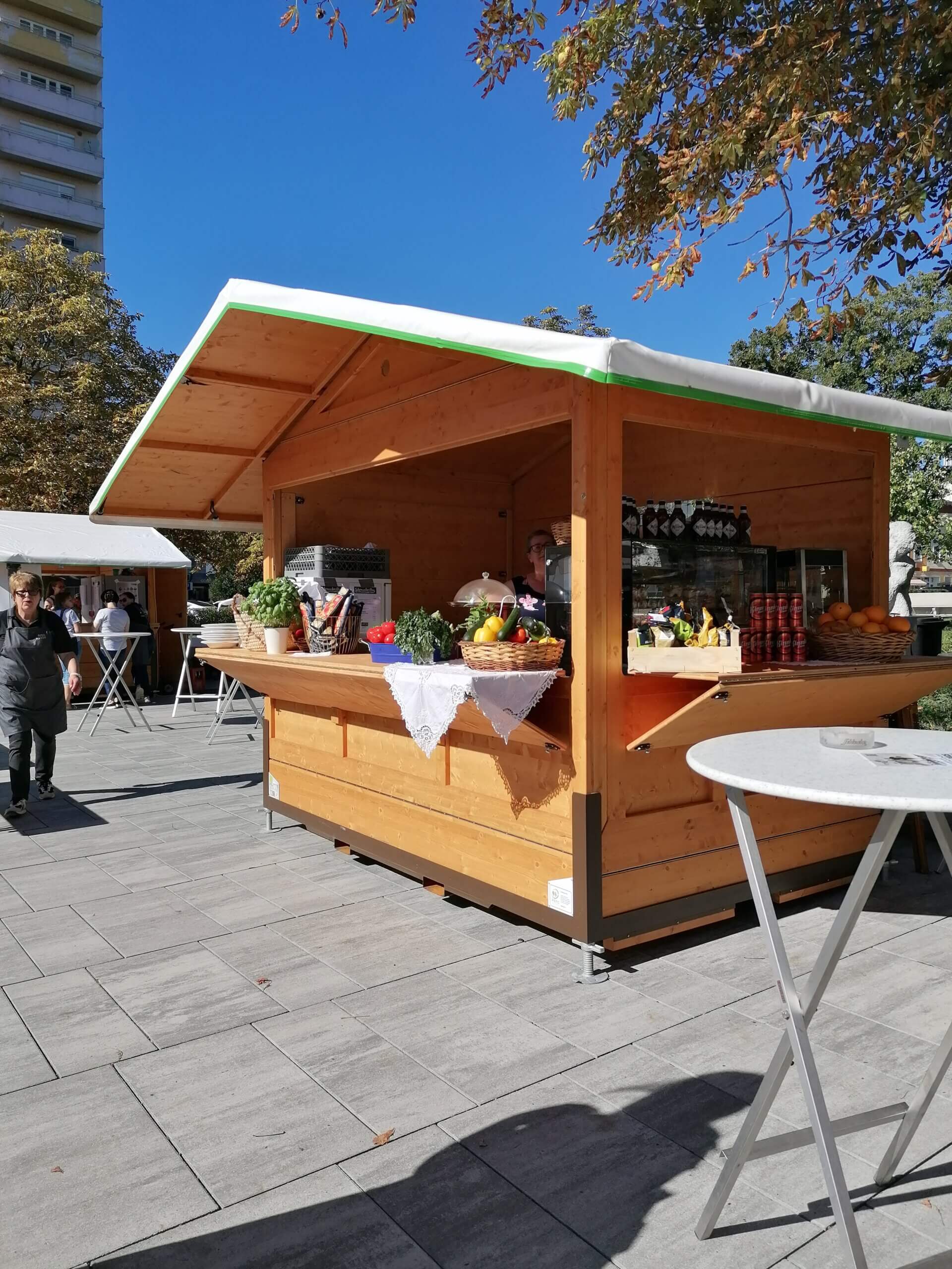 Holzverkaufsstand auf Marktplatz mit frischem Obst und Kräutern, blauer Himmel, Herbstbäume im Hintergrund.