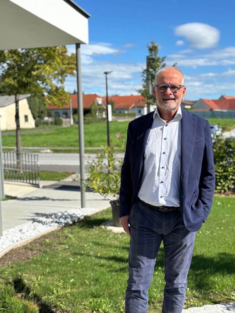 Ein Mann im Anzug steht draußen bei sonnigem Wetter vor einem modernen Gebäude mit Garten im Hintergrund.