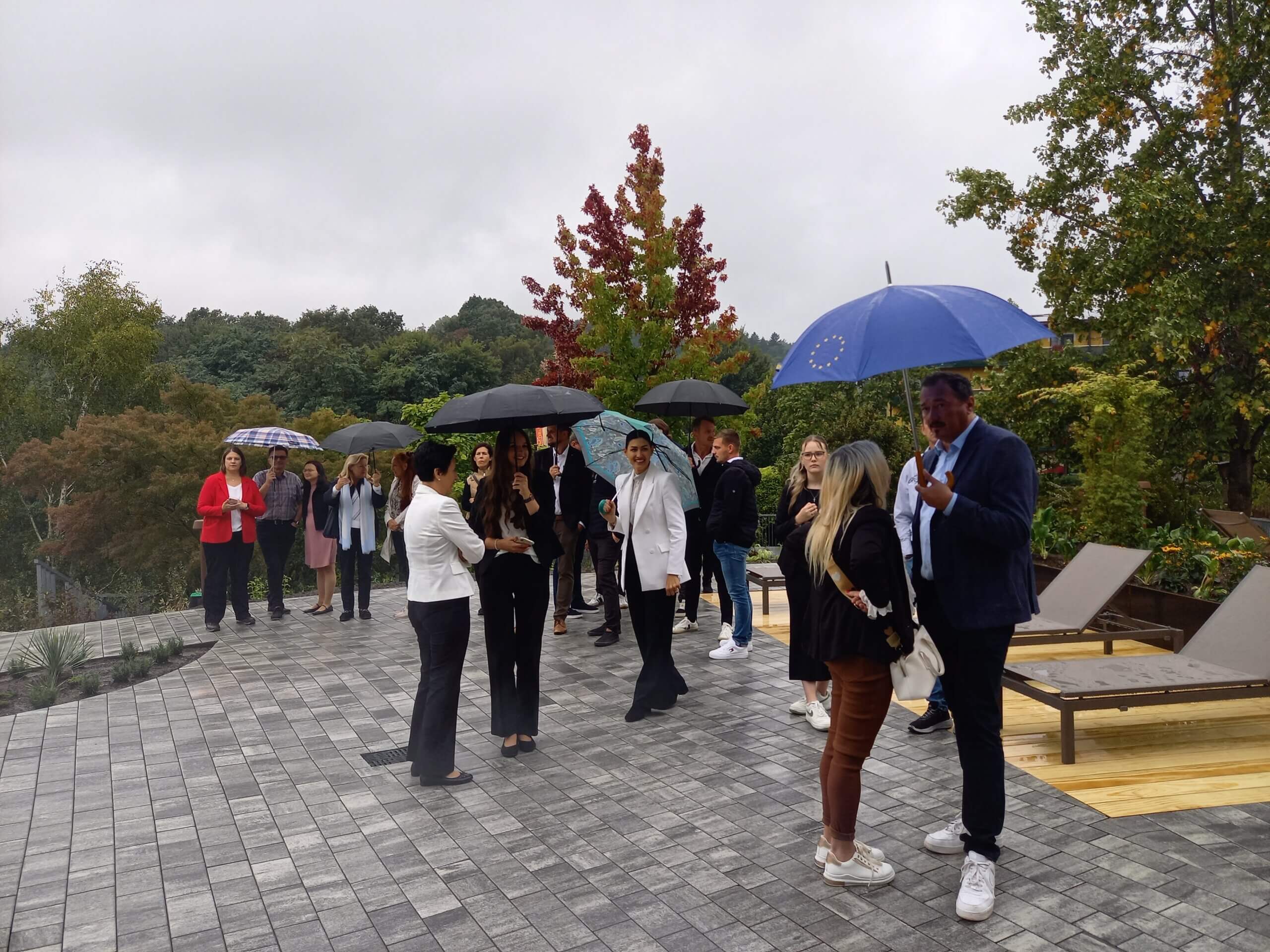 Gruppe von Menschen mit Regenschirmen auf einer Terrasse bei bedecktem Himmel, umgeben von herbstlichen Bäumen.