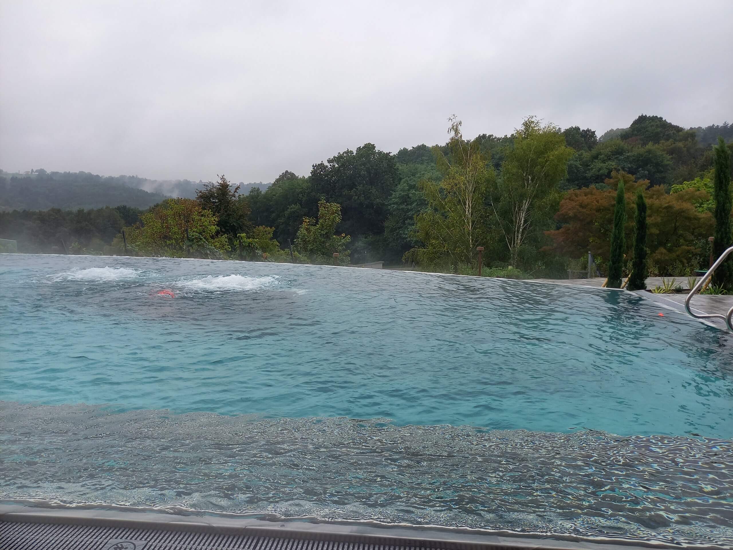 Infinity-Pool mit Dampfeffekt und Aussicht auf bewaldete Hügel unter grauem Himmel. Entspannende Naturkulisse.