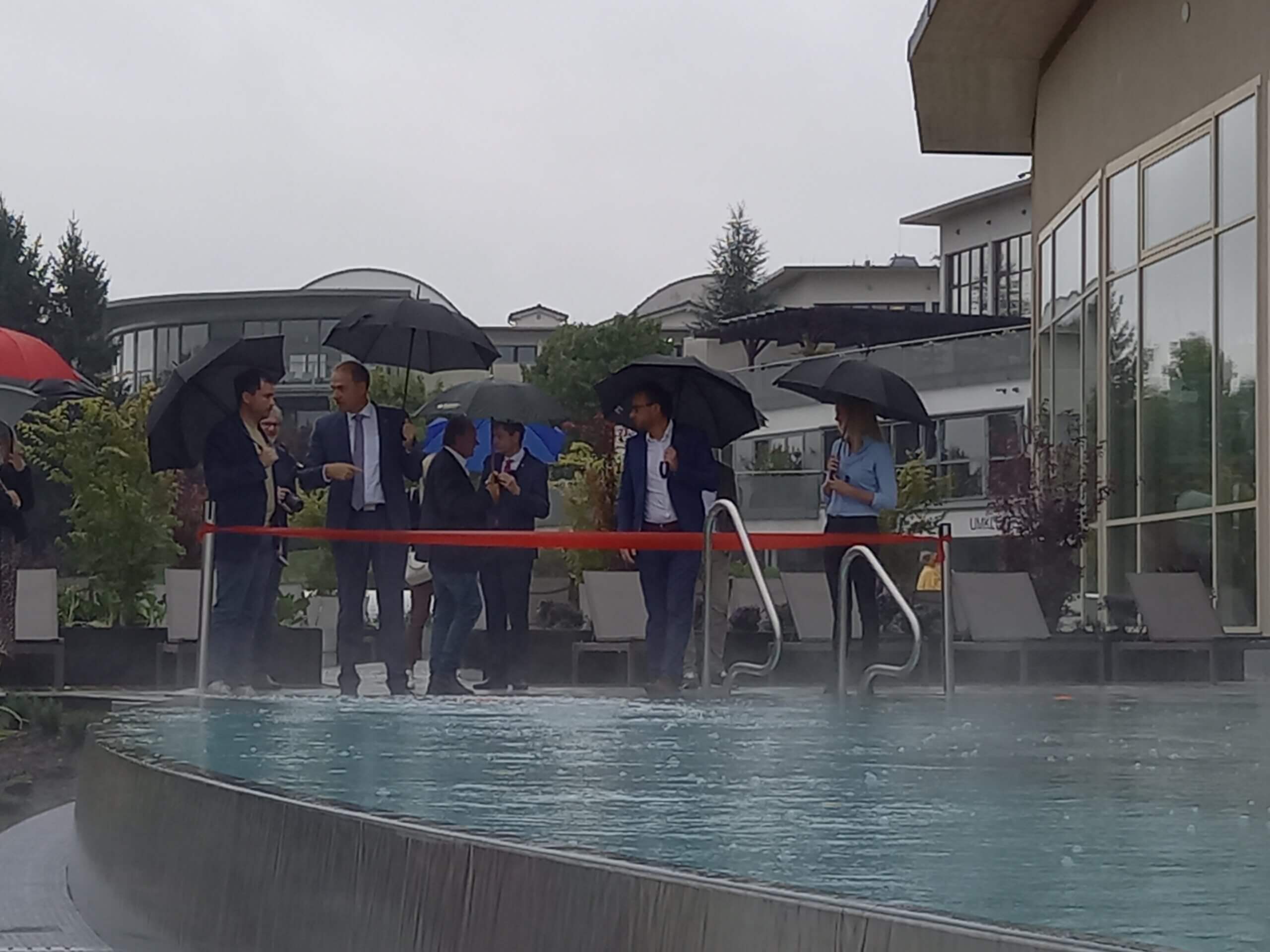 Gruppeneröffnung eines Schwimmbads bei Regenwetter, Personen mit Regenschirmen stehen vor dem Pool hinter einem roten Band.