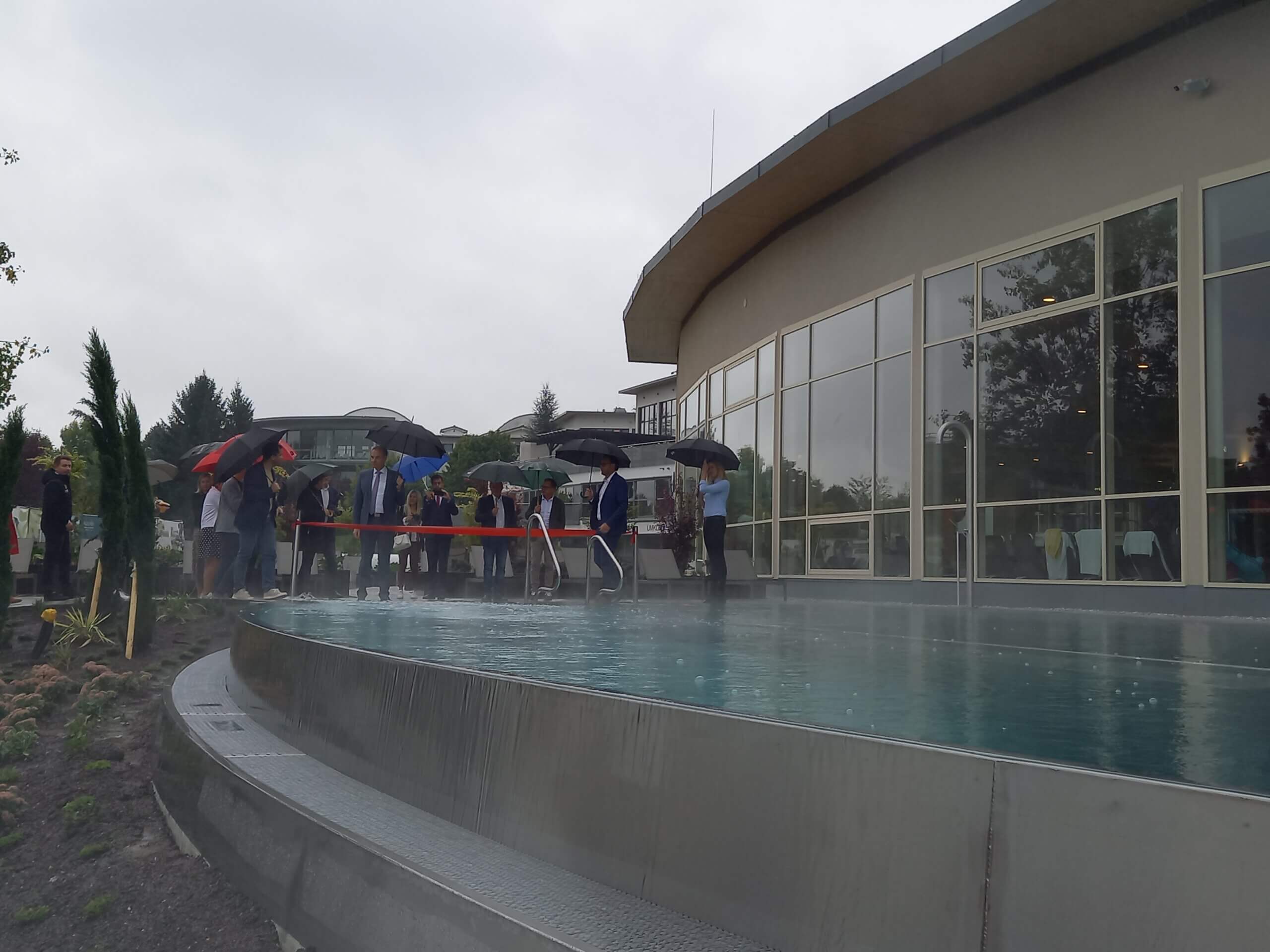 Menschen mit Regenschirmen vor einem modernen Gebäude bei einer Veranstaltung neben einem Pool.