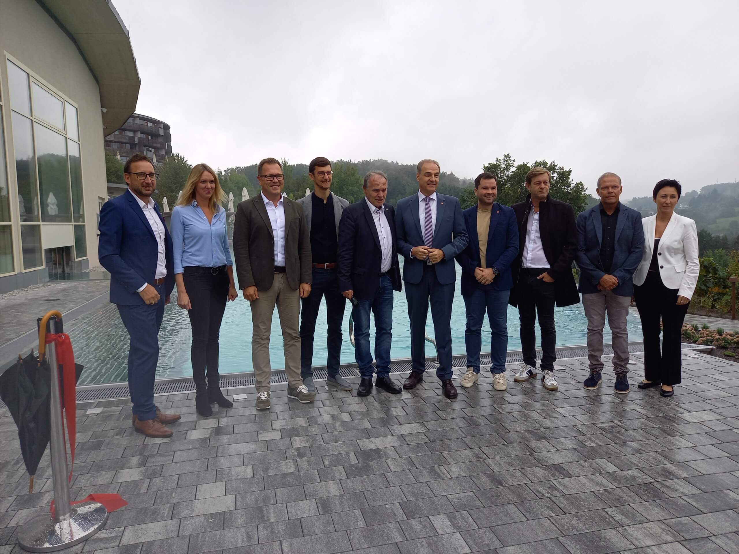 Gruppe von zehn Personen in Businesskleidung posiert draußen vor einem Pool an einem bewölkten Tag.