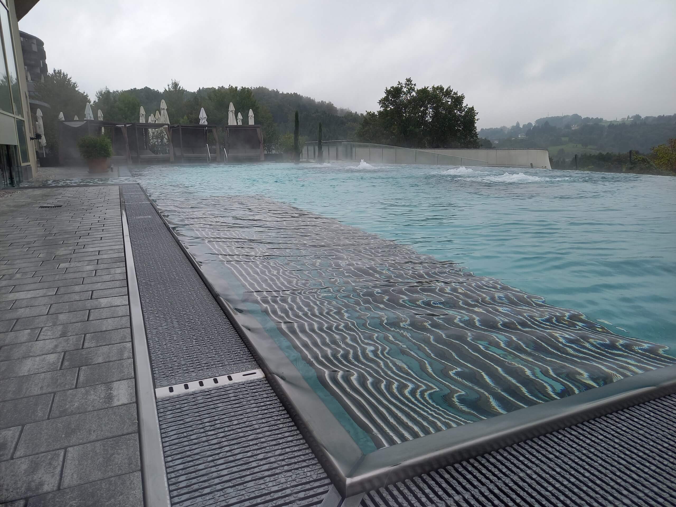 Infinity-Pool im Freien mit Aussicht auf bewaldete Hügel und grauem Himmel, ideal für Entspannung und Erholung.