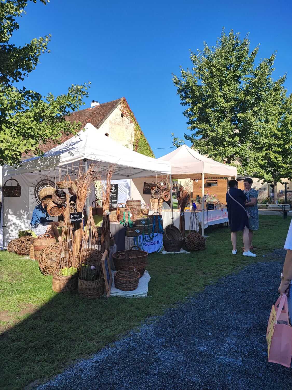 Marktstand mit geflochtenen Körben unter weißen Pavillons, Sonnenschein, Bäume im Hintergrund. Perfekt für Outdoor-Einkäufe.