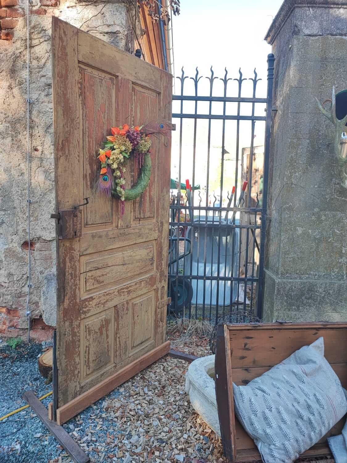 Historische Holztür mit herbstlichem Kranz vor schmiedeeisernem Tor in rustikaler Umgebung.