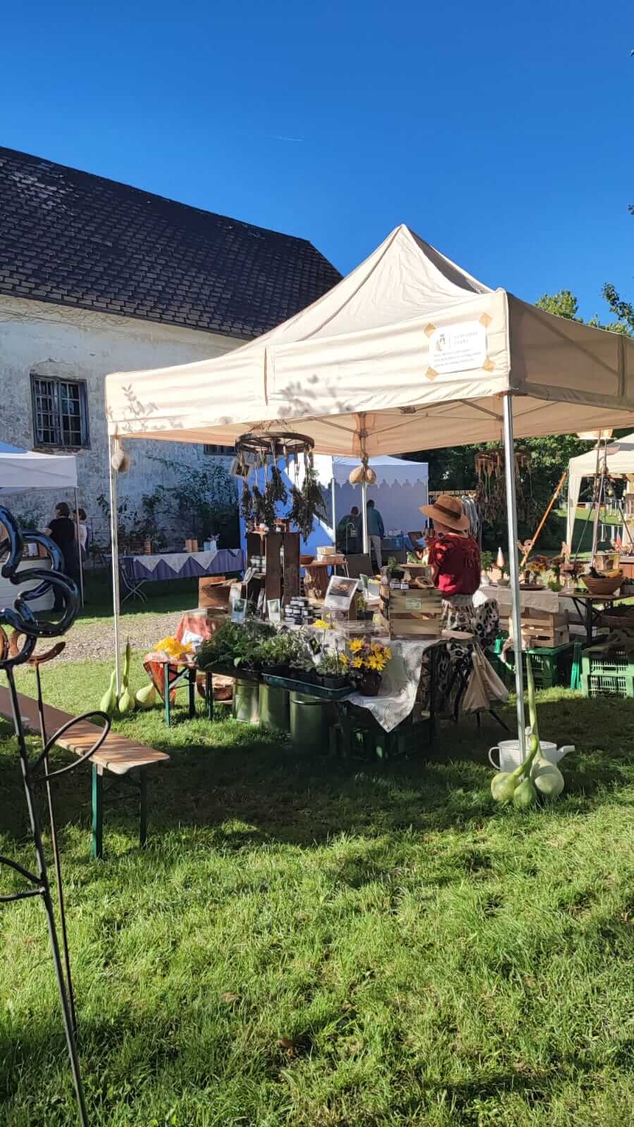 Marktstand im Freien mit Pflanzen und Produkten unter einem Zelt bei sonnigem Wetter vor einem alten Gebäude.