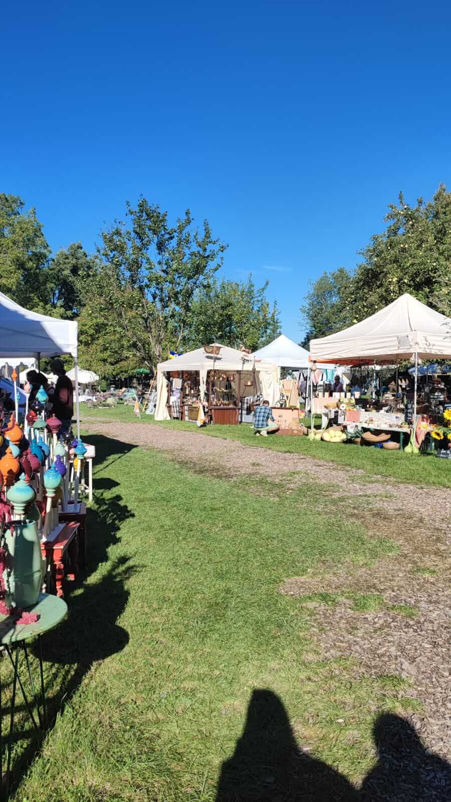 Freiluft-Kunsthandwerkermarkt mit bunten Ständen unter blauem Himmel, umgeben von grünen Bäumen.