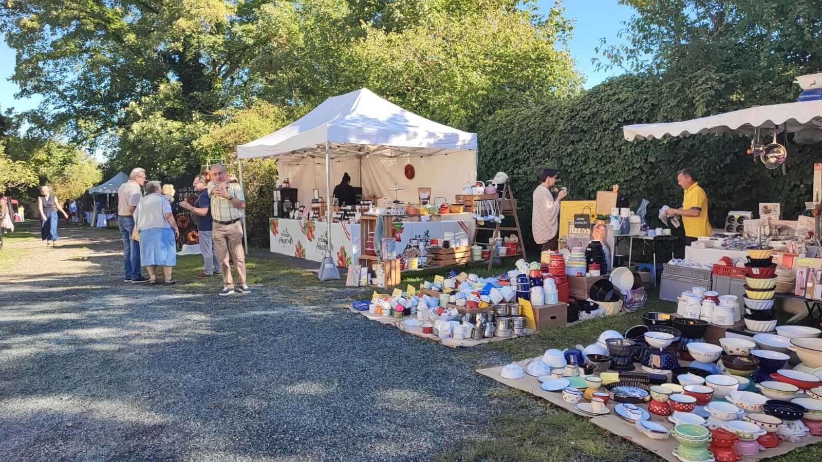 Menschen schlendern an Marktständen im Freien bei Sonnenschein vorbei, mit buntem Geschirr und Handwerkskunst.