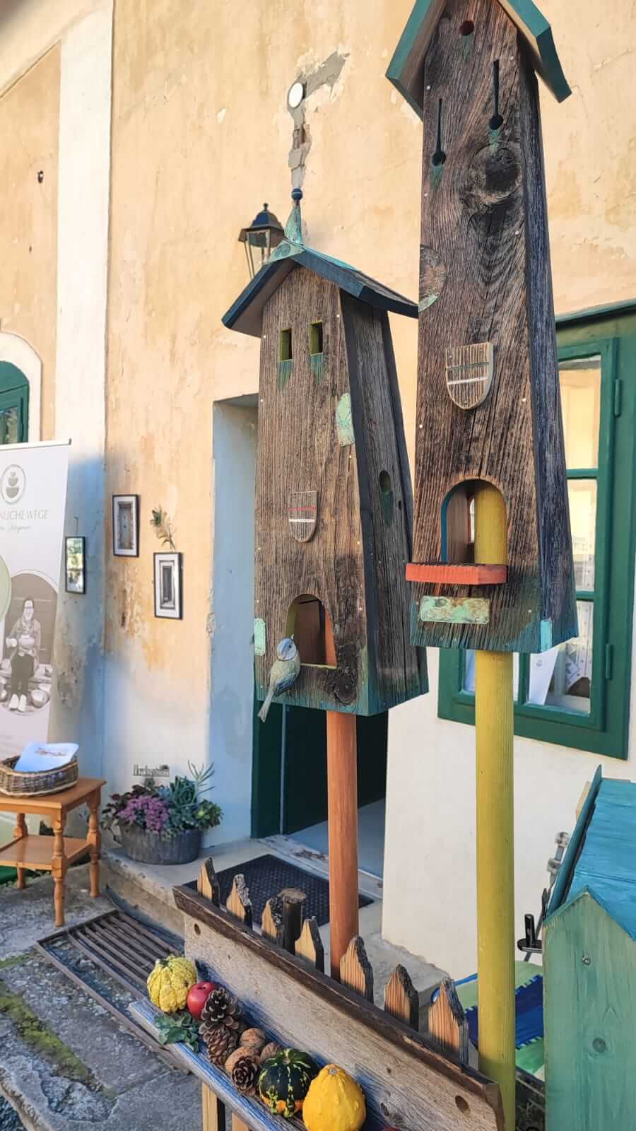 Rustikale Holzvogelhäuser mit dekorativen Kürbissen und Kiefernzapfen vor einem alten Gebäude.