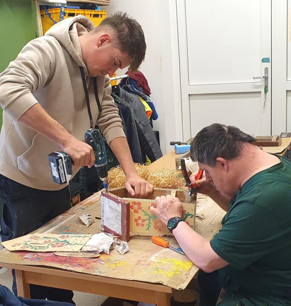 Zwei Personen beim Basteln: Eine nutzt eine Bohrmaschine, die andere werkelt an einer dekorierten Holzbox im Werkraum.