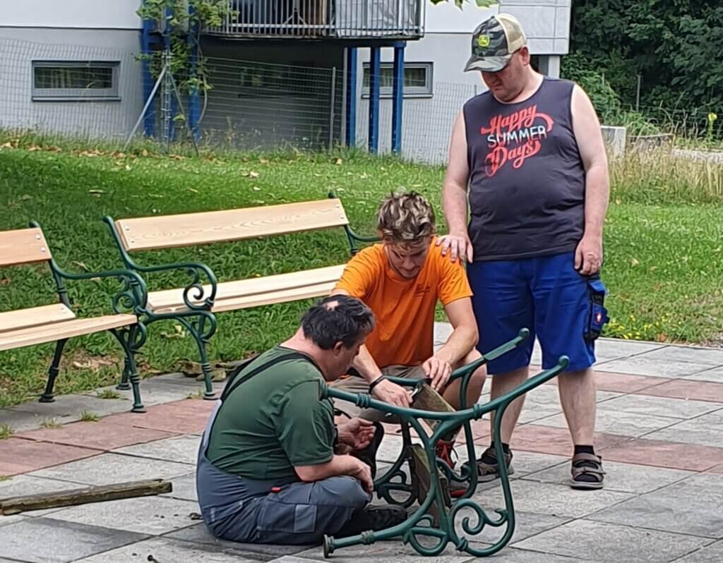 Drei Männer reparieren eine Parkbank im Freien. Zusammenarbeit und handwerkliche Arbeit in einer grünen Umgebung.