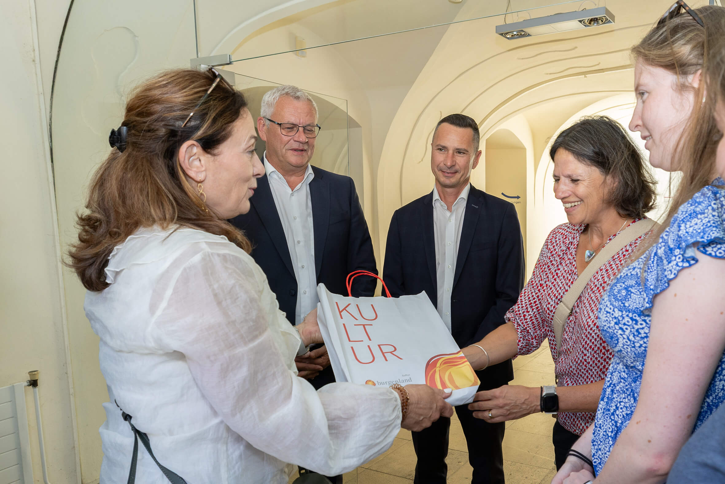 Personen tauschen Geschenke bei einer Kulturveranstaltung aus, lächeln und freuen sich im Gebäude mit weißem Bogen.