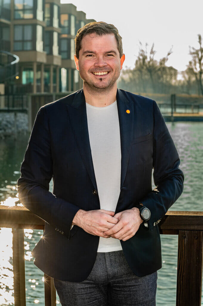 Ein Mann in einem blauen Sakko lächelt vor einer modernen Gebäude- und Wasserhintergrundkulisse.