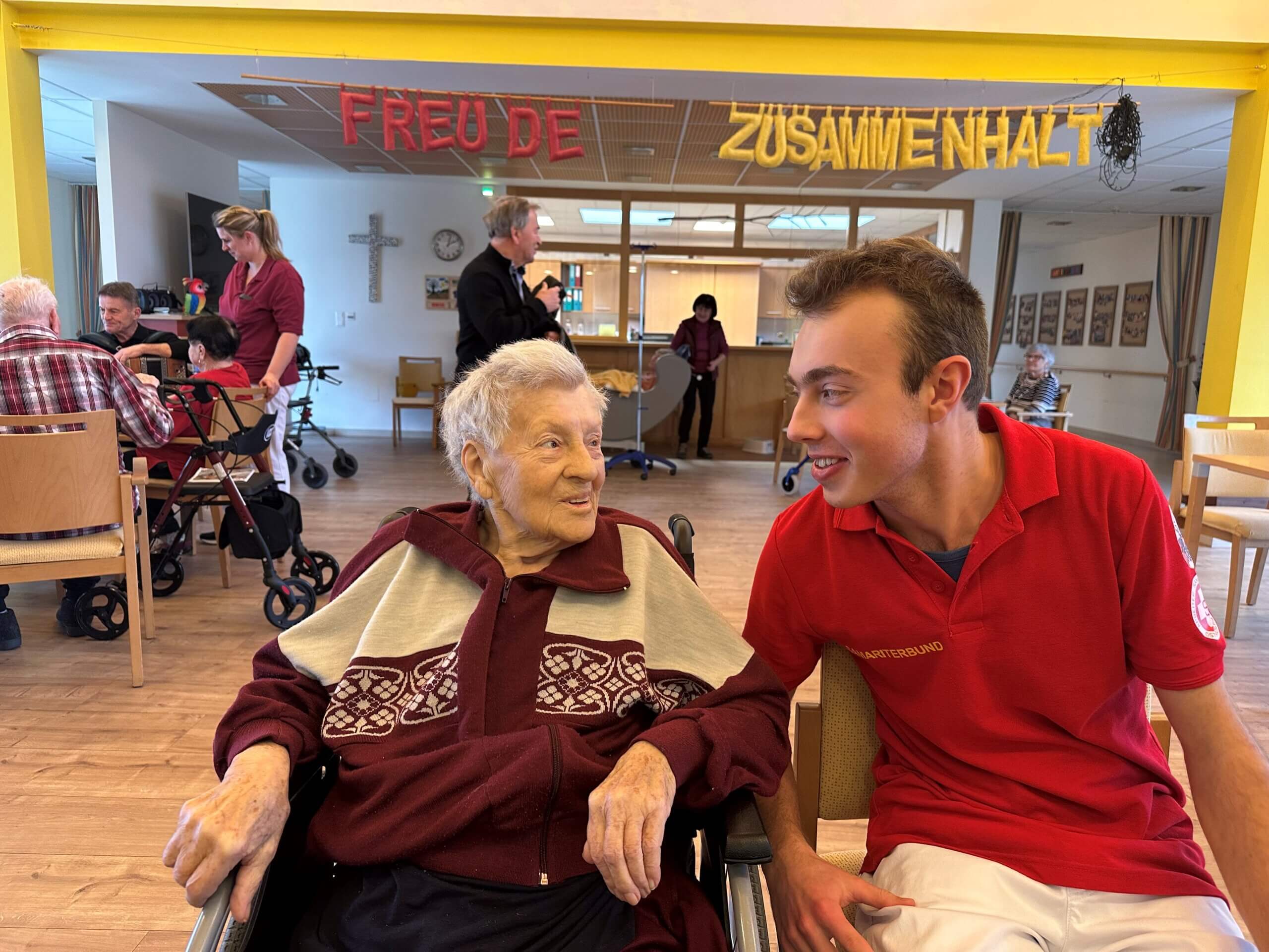 Pflegekraft unterhält sich mit Seniorin im Gemeinschaftsraum eines Pflegeheims, unter dem Motto „Freude und Zusammenhalt“.