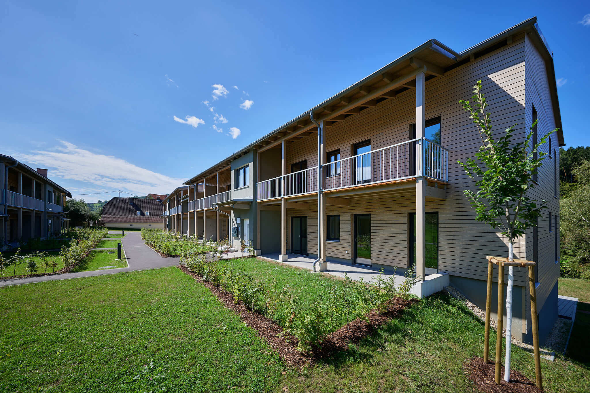Moderne Wohnanlage mit Holzfassade und gepflegter Gartenanlage unter blauem Himmel.