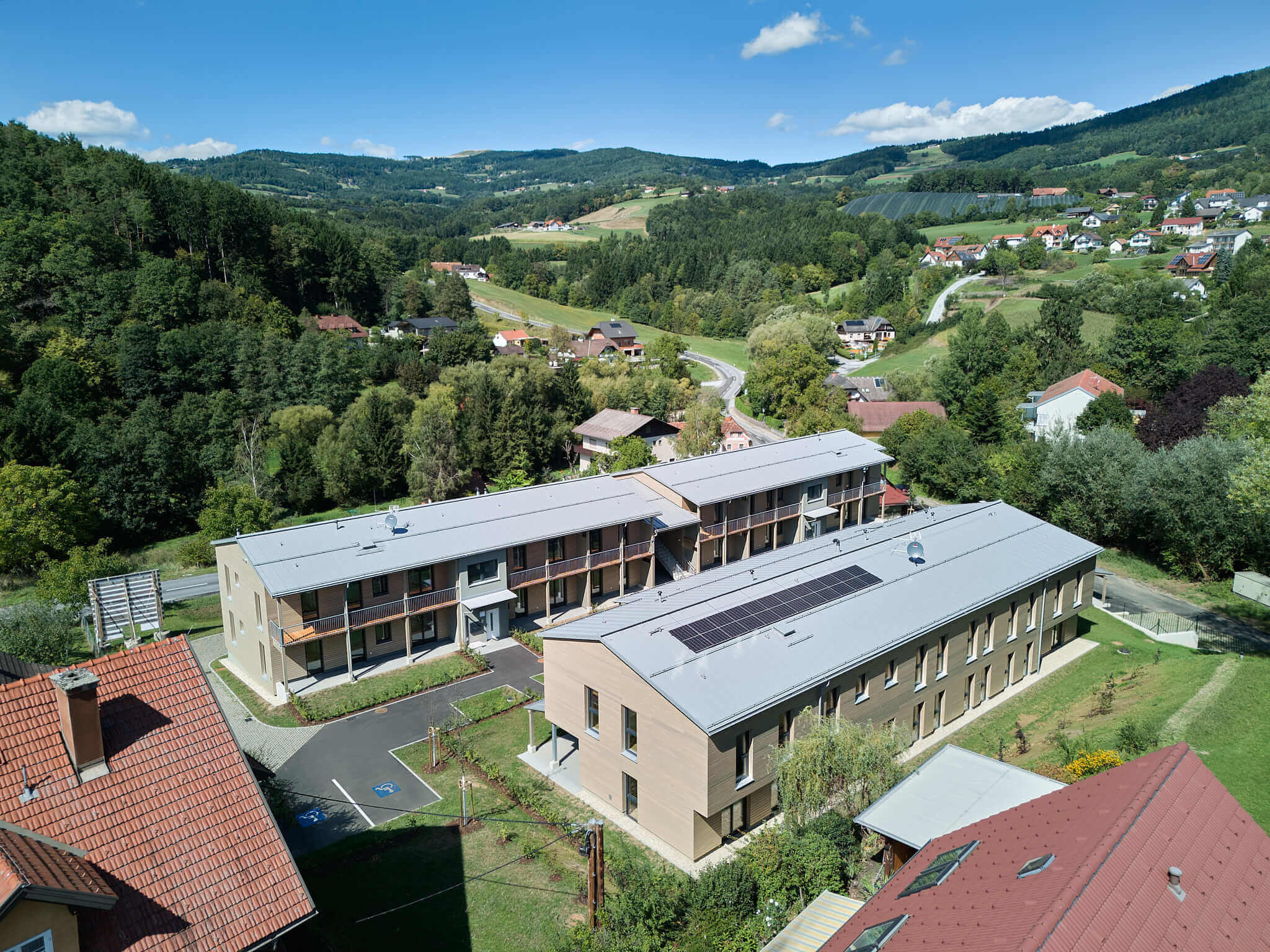 Moderne Wohnanlage mit Solarpanels in idyllischer Landschaft, umgeben von Wald und Hügeln, sonniger Tag.