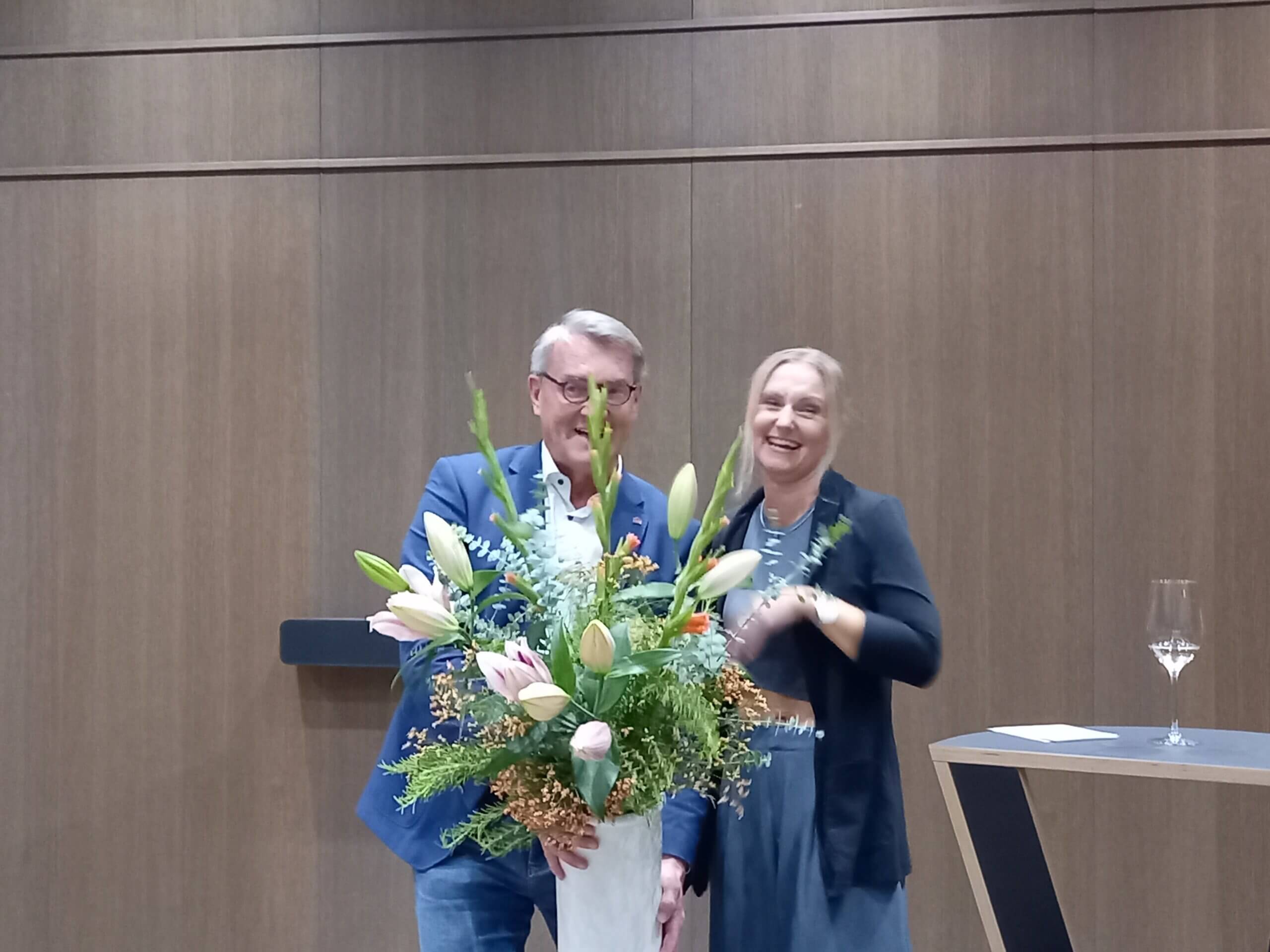 Mann und Frau mit großem Blumenstrauß lächeln vor Holzverkleidung bei einer Veranstaltung.