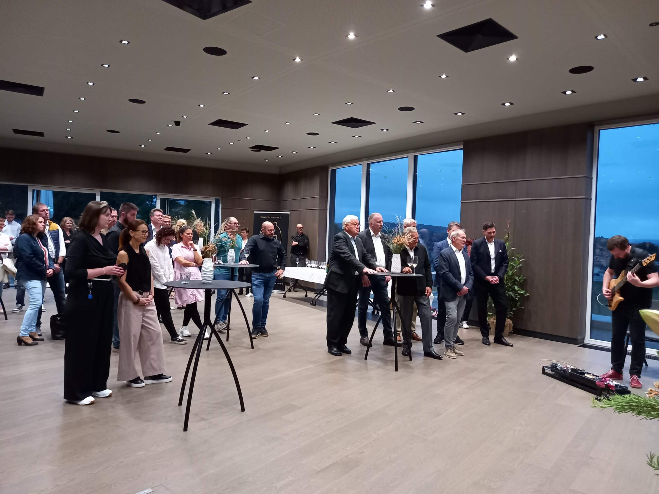 Gruppe von Menschen bei einer Veranstaltung mit Livemusik in modernem Saal, Musiker spielt Gitarre, Publikum hört zu.