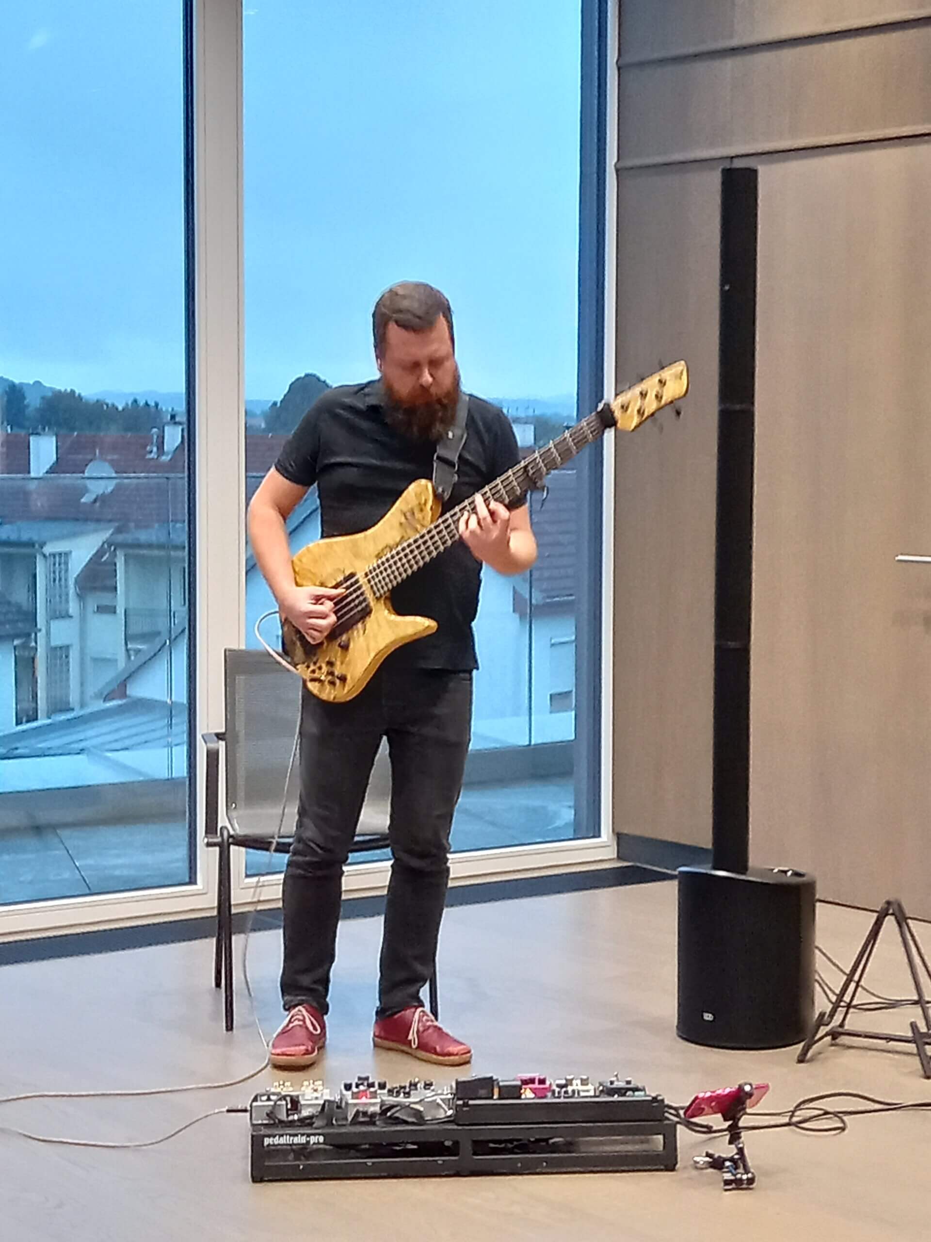 Musiker spielt E-Gitarre vor Fenster mit Effektpedalen, konzentriert bei einem Auftritt in modernem Raum.