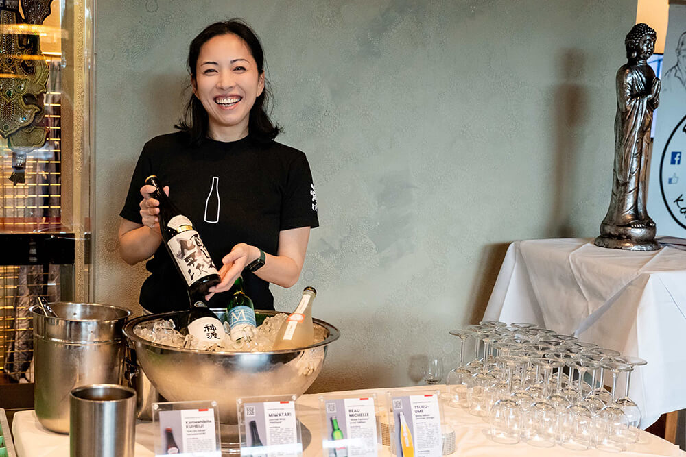 Frau präsentiert Sake-Varianten auf einer Veranstaltung, lächelnd mit mehreren Flaschen in einer Schale voller Eiswürfel.