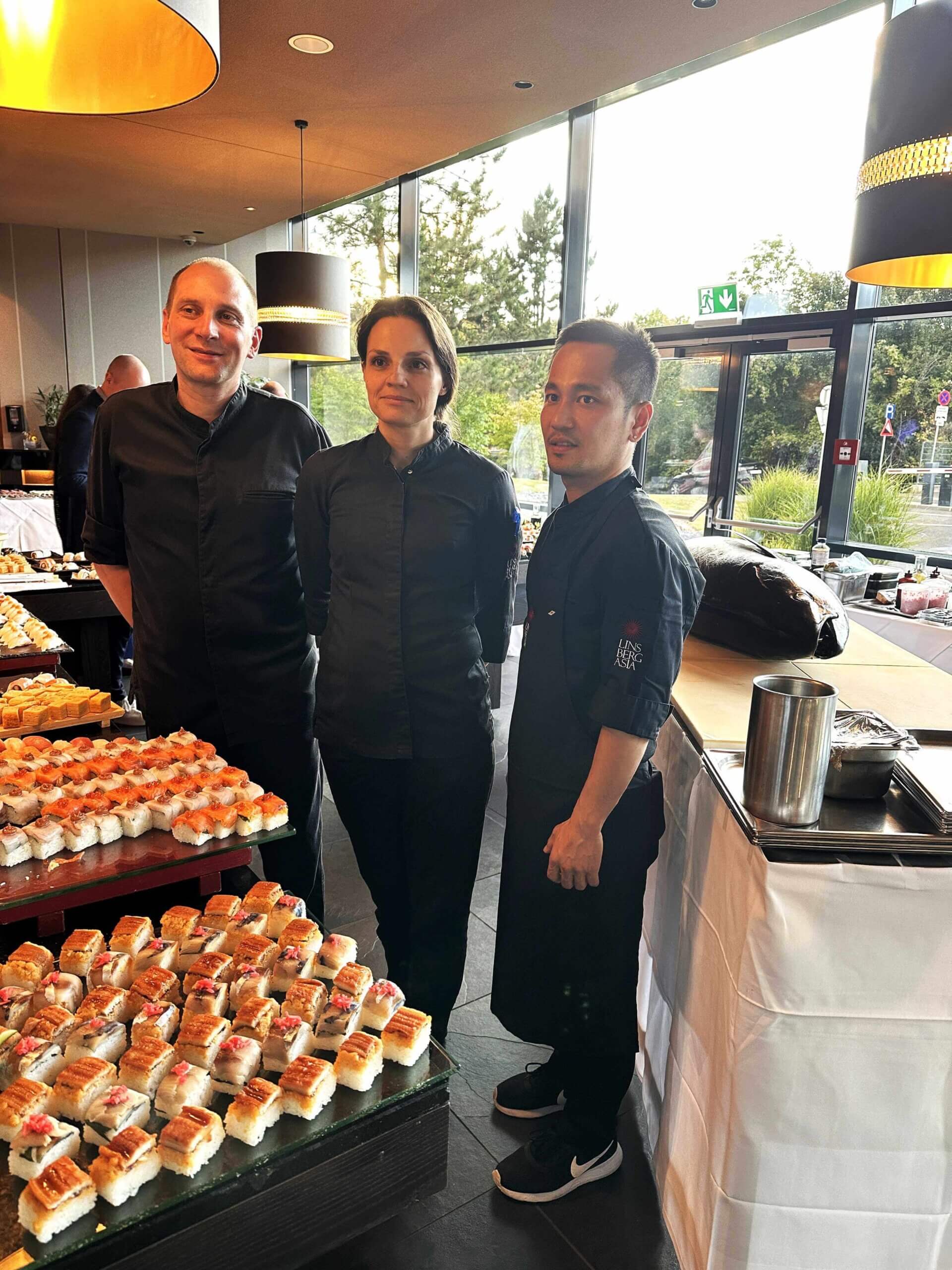 Köche präsentieren Buffet mit Sushi-Kreationen in modernem Restaurant mit großen Fenstern im Hintergrund.