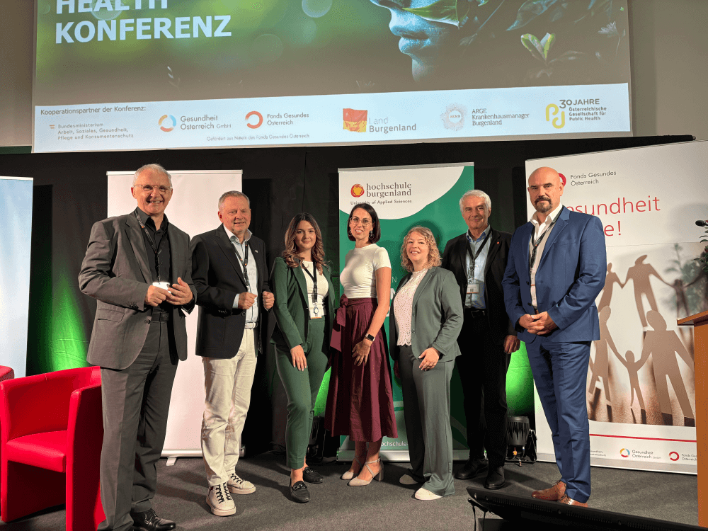 Gruppe von Menschen auf der Gesundheitskonferenz in Österreich mit Logos von Partnern im Hintergrund.